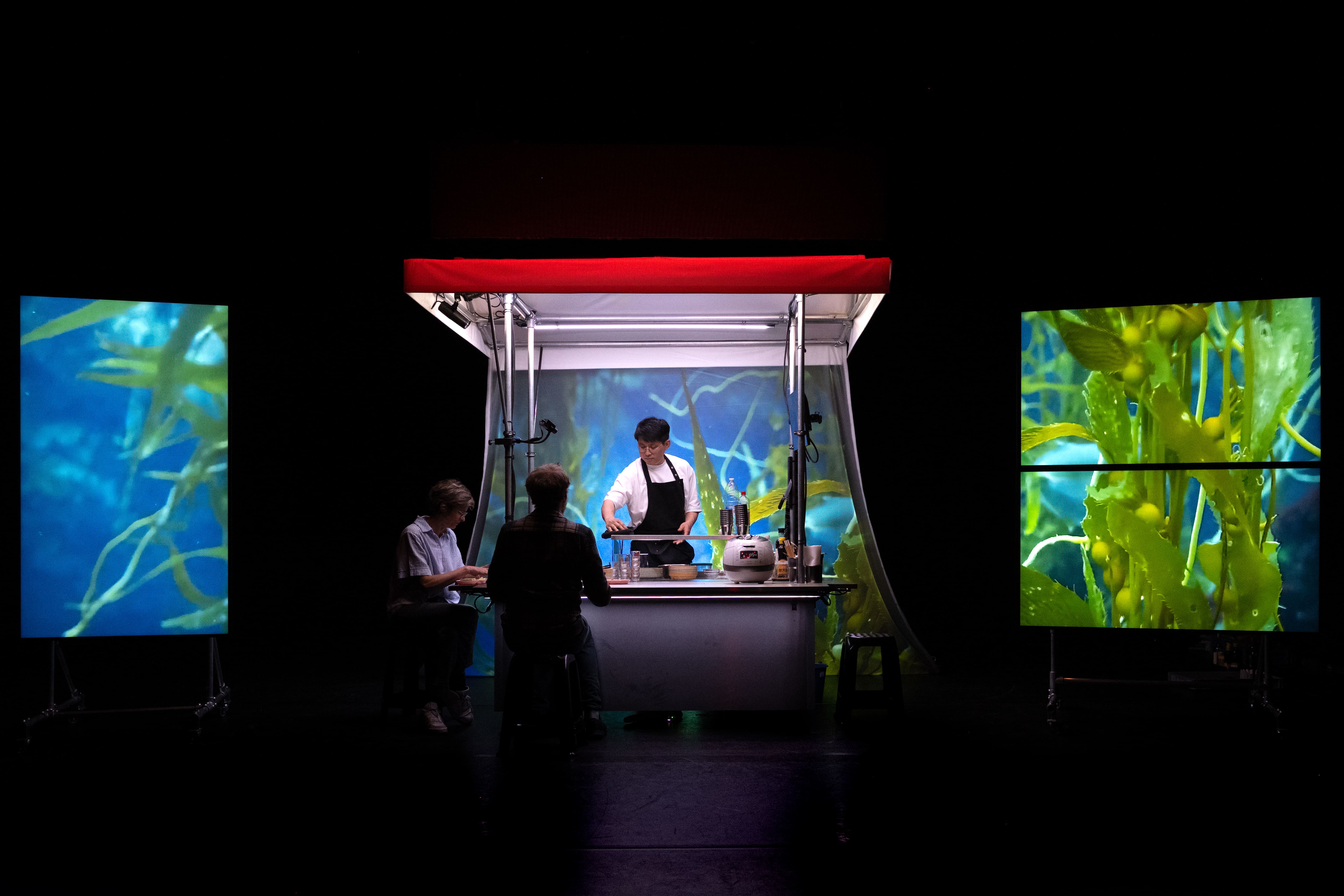 © Bea Borgers A stage art installation, showing a lit Korean food stall with a chef and two seated customers. The background of the stall and two large side LED screens display vibrant underwater images of green and blue kelp, contrasting with the dark stage and the stall's red awning. - Bea Borgers
