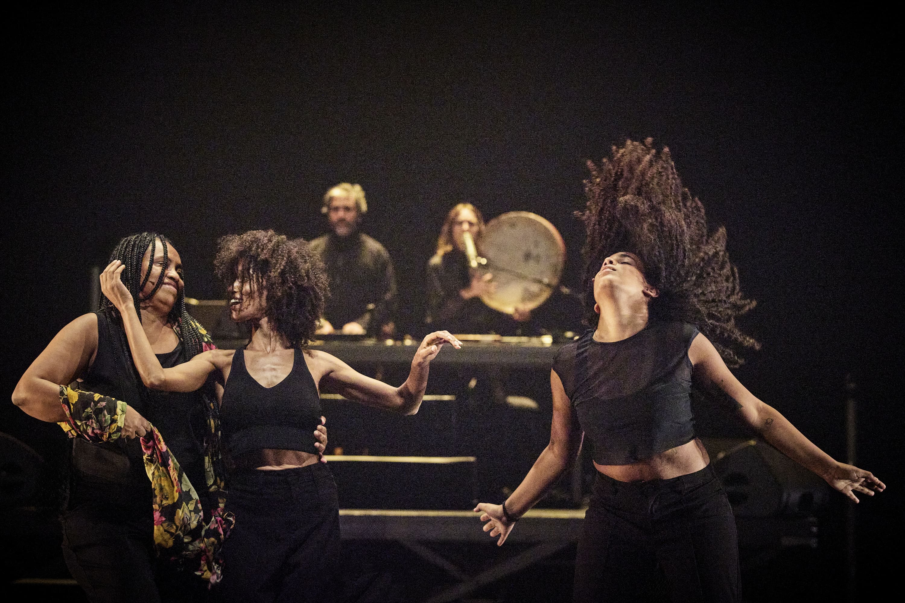 Três mulheres dançam com energia em primeiro plano, enquanto dois músicos atuam no palco, ao fundo. - Christophe Raynaud De Lage