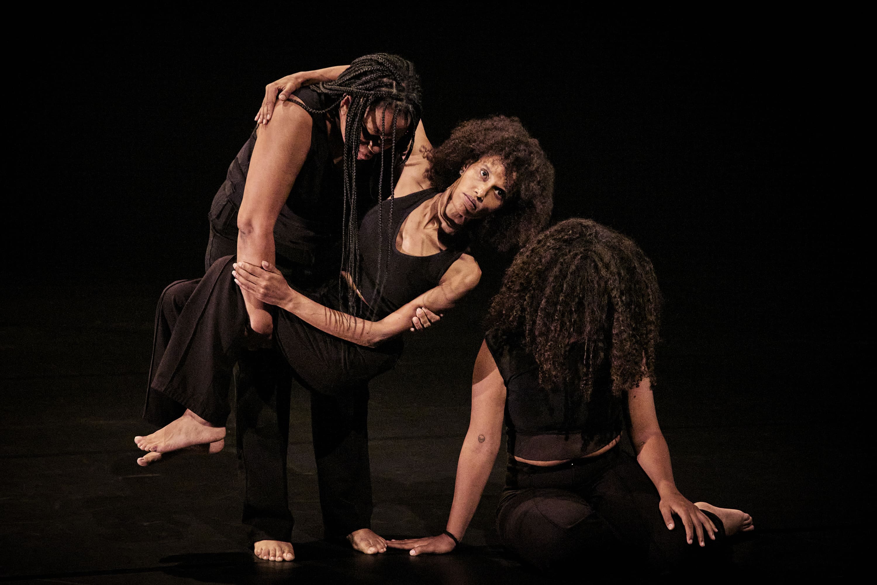 Três mulheres negras com roupas pretas num palco escuro. No centro, uma mulher com cabelo afro está a ser amparada e levantada por outra mulher com tranças. A terceira mulher, com cabelo encaracolado, está sentada no chão, em primeiro plano, com o rosto tapado pelo cabelo, apoiando-se nas mãos. A composição sugere apoio mútuo e desamparo. - Christophe Raynaud De Lage