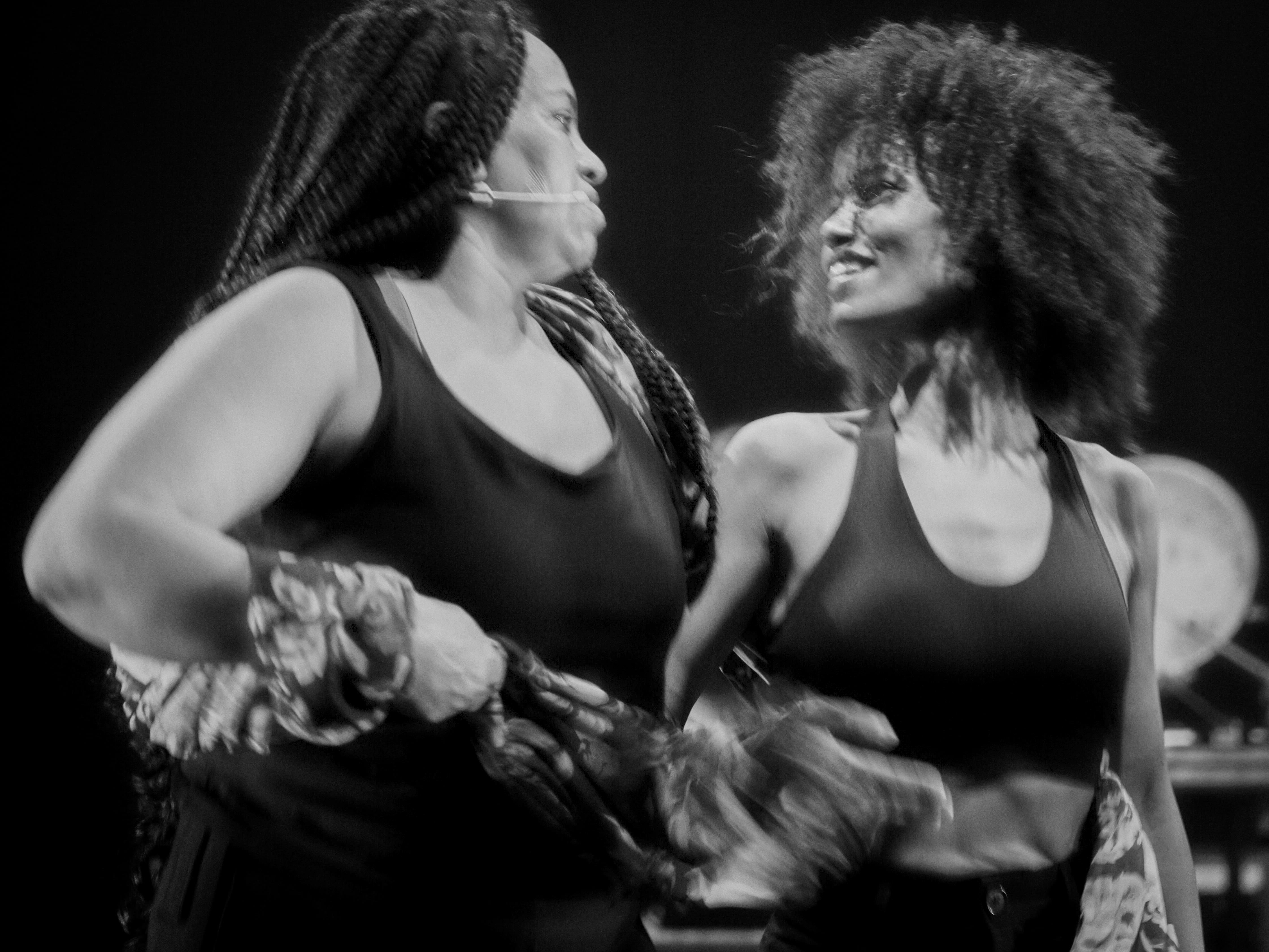 Uma fotografia a preto e branco em plano médio mostra duas mulheres negras num palco, olhando-se intensamente e sorrindo uma para a outra. Ambas vestem tops pretos. A mulher à esquerda tem tranças e um microfone de cabeça, e segura um tecido estampado nas mãos. A mulher à direita tem cabelo afro e usa um top de alças. O fundo está escuro e desfocado. - Lea Skayem