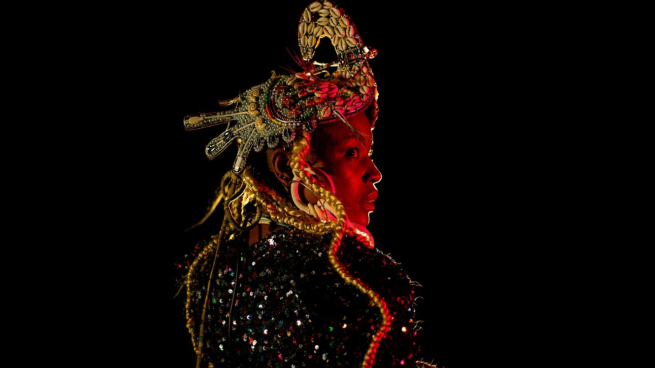 ALKANTARA - Retrato em plano aproximado de perfil de uma mulher negra iluminada por uma luz avermelhada sobre fundo preto. Ela usa um adereço de cabeça elaborado com búzios e detalhes metálicos, tranças loiras compridas e uma peça de roupa brilhante com lantejoulas. - ©Carolina Maya