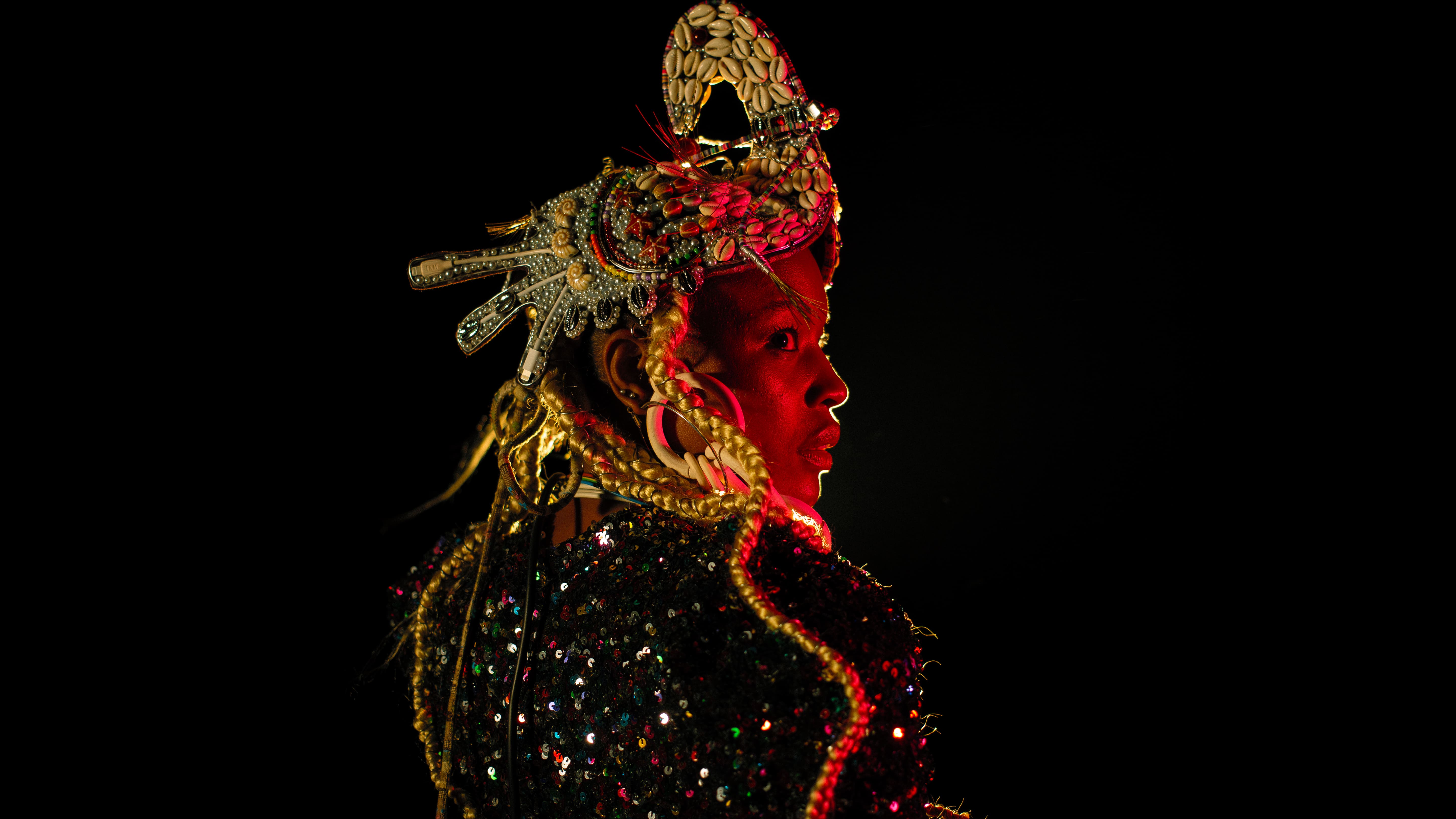 Retrato em plano aproximado de perfil de uma mulher negra iluminada por uma luz avermelhada sobre fundo preto. Ela usa um adereço de cabeça elaborado com búzios e detalhes metálicos, tranças loiras compridas e uma peça de roupa brilhante com lantejoulas. - Carolina Maya