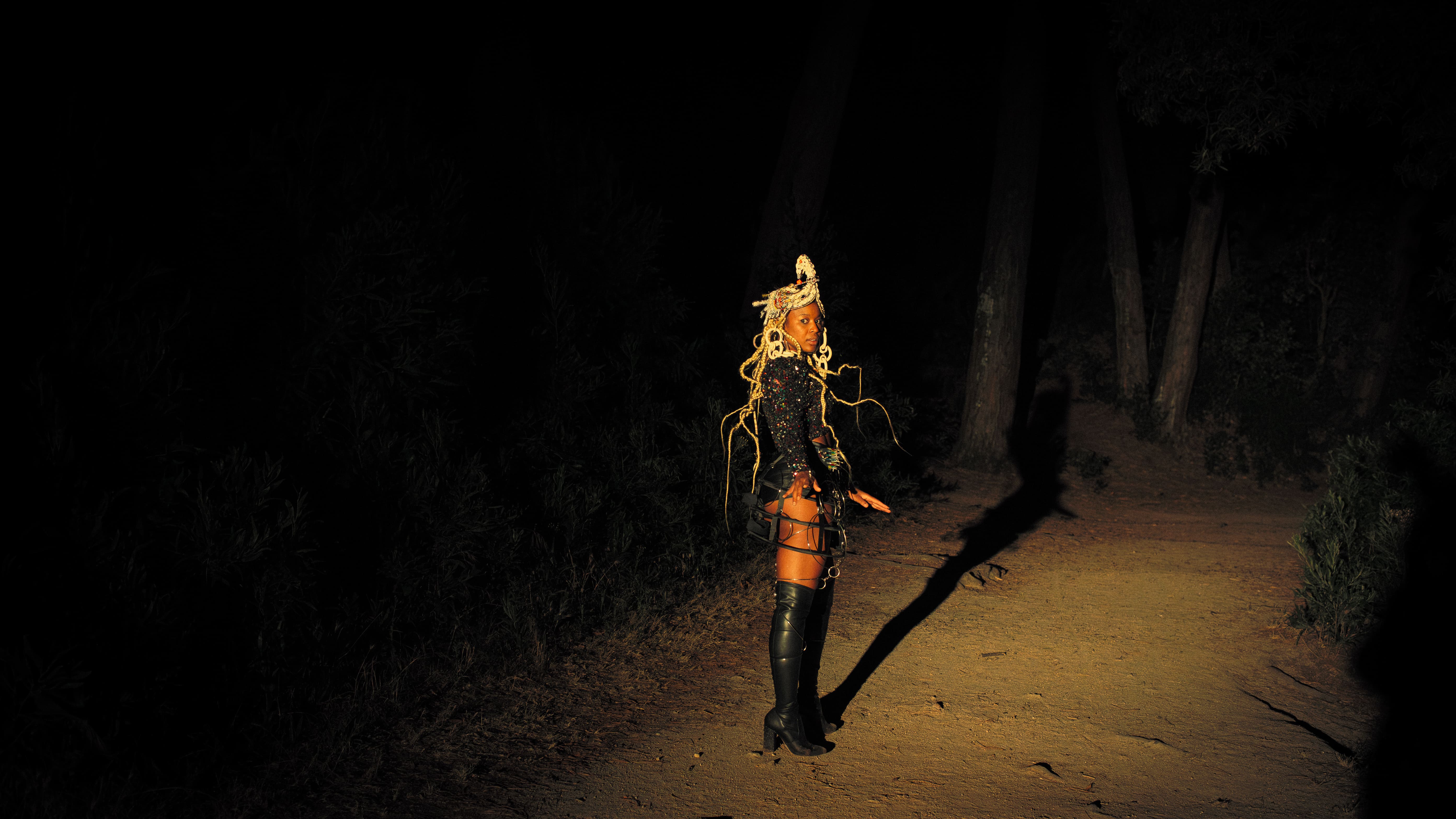 Figura inteira de uma mulher negra parada num caminho de terra à noite, cercada pela escuridão de uma floresta. Está iluminada por um foco de luz amarela, vestindo um fato com lantejoulas brilhantes, botas pretas de cano alto acima do joelho, adereço de cabeça com búzios e longas tranças soltas. - Carolina Maya