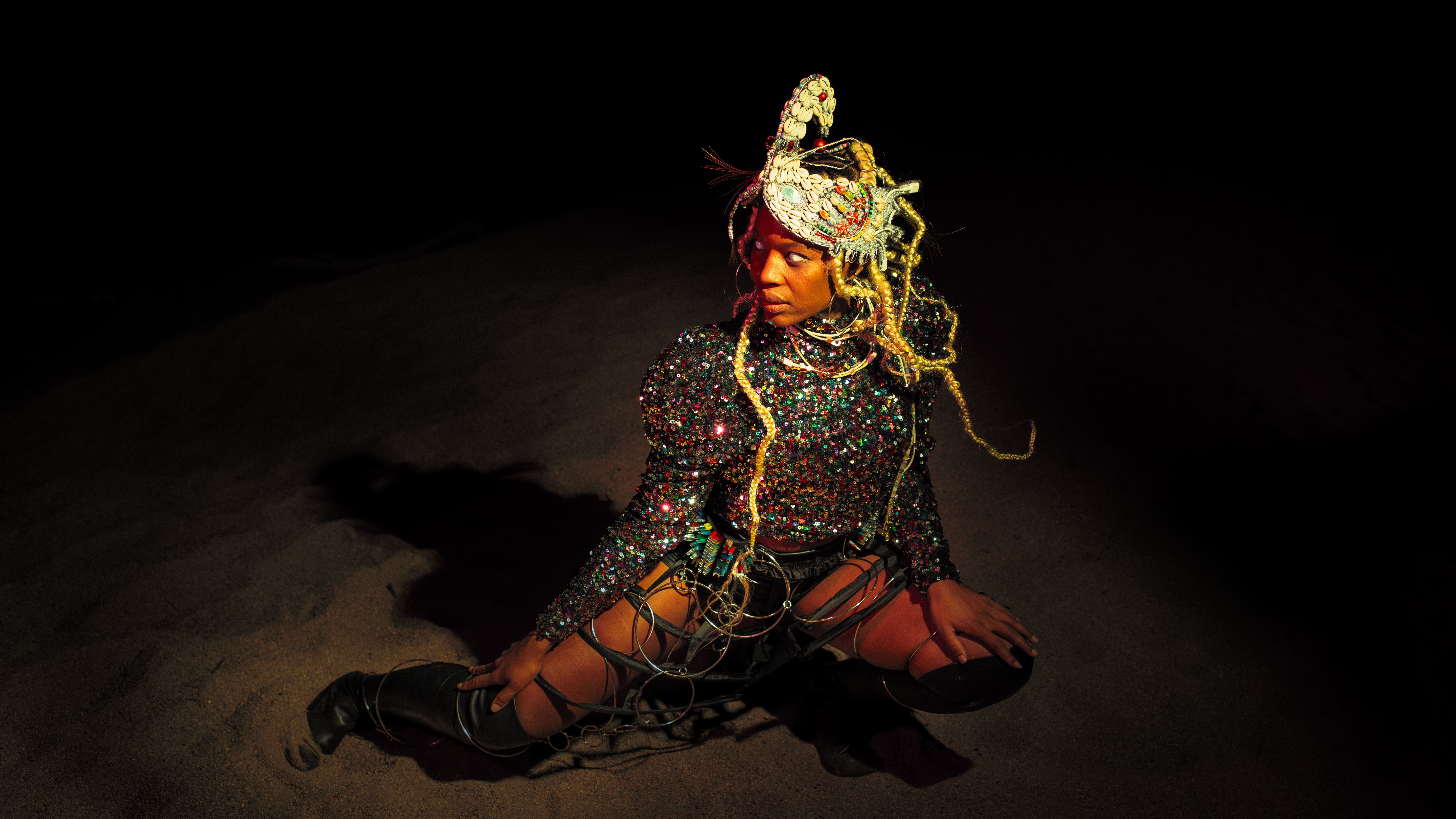 Plano médio de uma mulher negra ajoelhada na areia no meio da escuridão, sob um foco de luz quente. Usa um topo brilhante de lantejoulas, botas pretas de cano alto, uma estrutura metálica nas pernas e um ornamento de cabeça detalhado com búzios e tranças loiras. - Carolina Maya