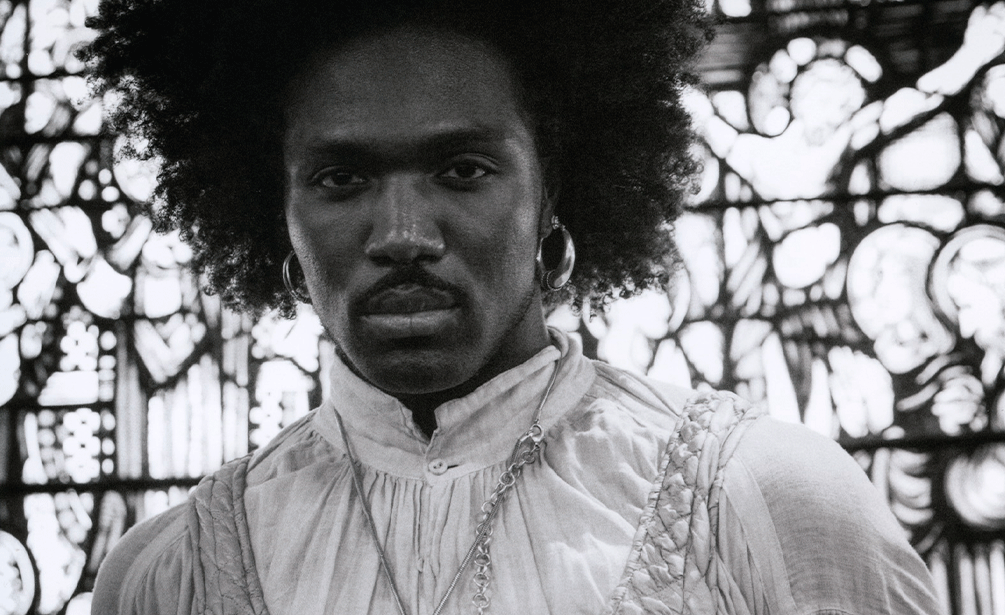 © Geoffrey Baptiste Retrato a preto e branco de um homem negro de frente, com cabelo afro, bigode e argolas nas orelhas, olhando diretamente para a câmara. Veste uma camisa clara com pregas e gola subida e correntes ao pescoço, posicionando-se à frente de um vitral trabalhado. - Geoffrey Baptiste