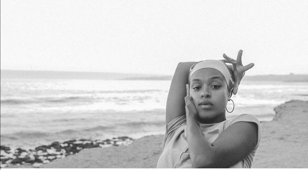 © Virginia Perez Retrato a preto e branco em plano médio de uma mulher jovem na praia com o mar ao fundo. Usa uma faixa/lenço na cabeça e argolas, posando com um braço erguido e a mão a emoldurar o rosto enquanto olha serenamente para a câmara. - Virginia Perez