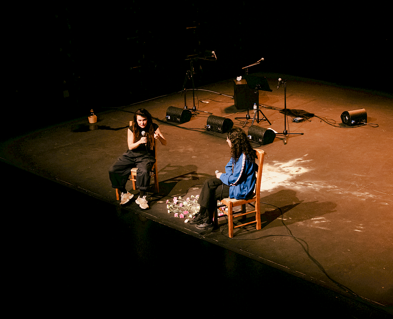 ALKANTARA - High-angle view of a dark stage where two people sit facing each other on wooden chairs, holding microphones during a talk. Pink flowers are scattered on the floor between them. The person on the left wears black trousers and a black t-shirt; the person on the right wears a blue sports jacket and dark trousers. Speakers and microphone stands are visible in the background. - ©Beatriz Pequeno