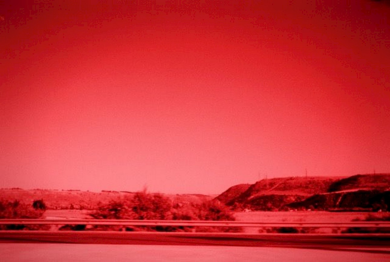 ALKANTARA - Paisagem de um sítio algures no campo, captada a partir de um veículo em movimento. Vemos um pouco da estrada e planícies. A fotografia está em tons de vermelho e preto. - ©Amel Malfait Lakhtara