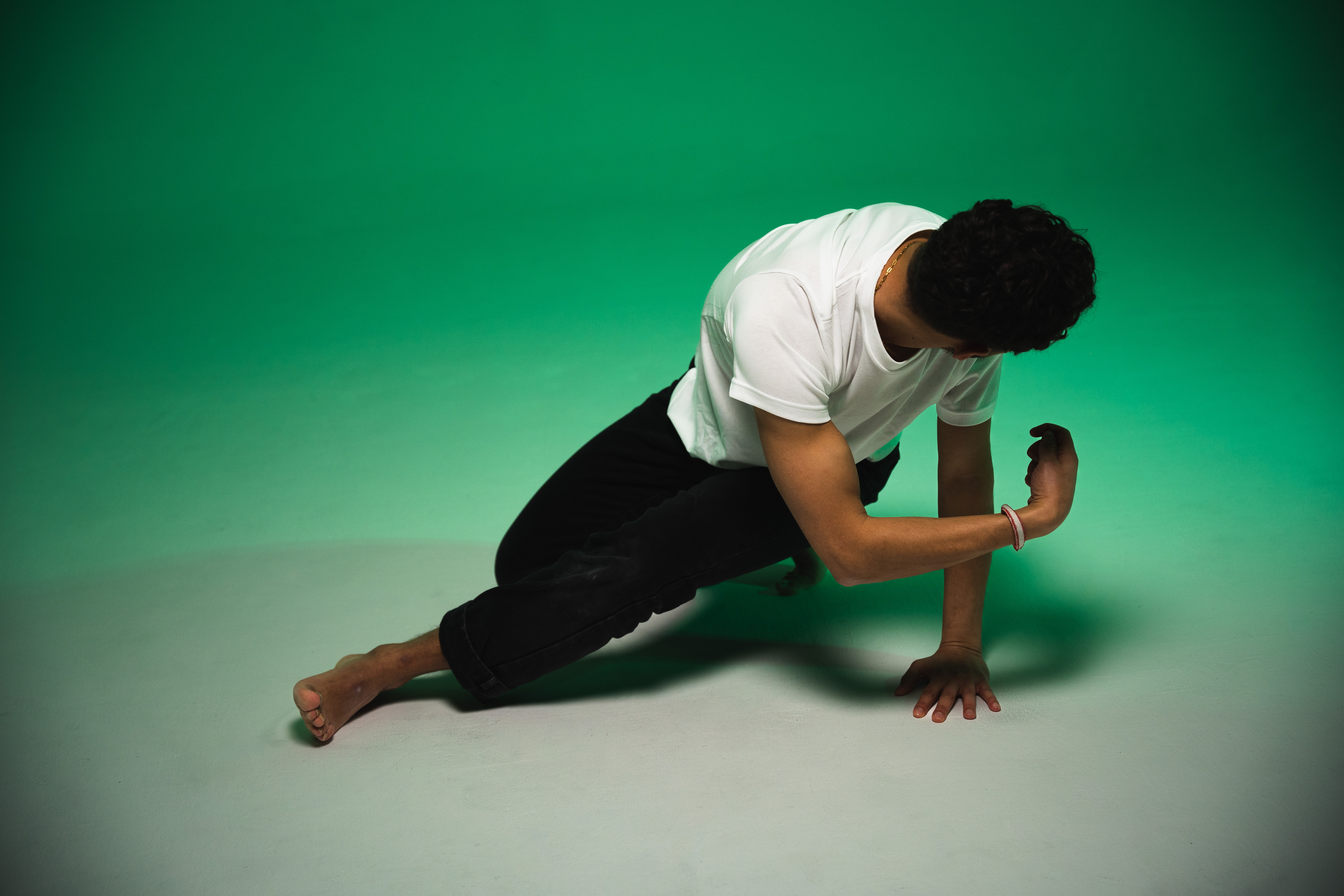 © Njaheut Valler Um homem descalço com t-shirt branca e calças pretas agachado num movimento de dança num estúdio com fundo verde. O corpo está inclinado para a frente com uma mão apoiada no chão e a cabeça virada para baixo. - Njaheut Valler