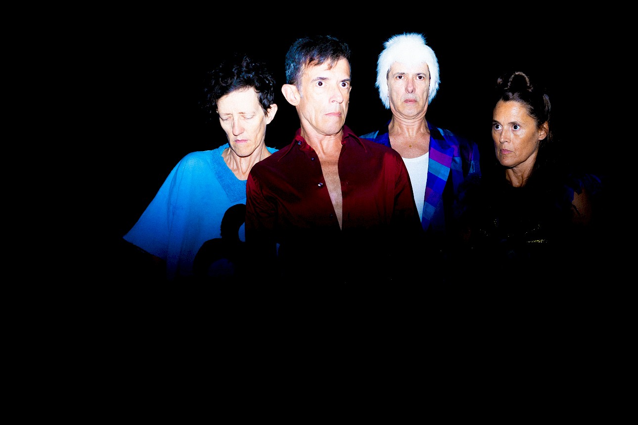 ALKANTARA - Four people pose lined up against a solid black background under dramatic flash lighting. The person on the left wears a light blue t-shirt with their eyes closed; the second wears an unbuttoned burgundy shirt; the third wears a short white wig and a blue and purple geometric patterned blazer; the person on the far right gazes intently towards the camera. - ©Alípio Padilha
