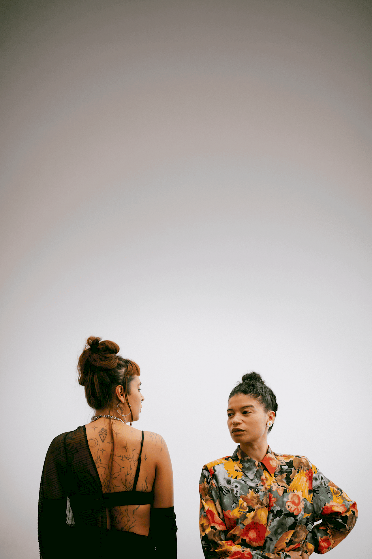 ALKANTARA - Two women in a studio against a plain light gray background. The woman on the left turns away showing back tattoos and a sheer black top; the woman on the right faces forward wearing a bright floral shirt.