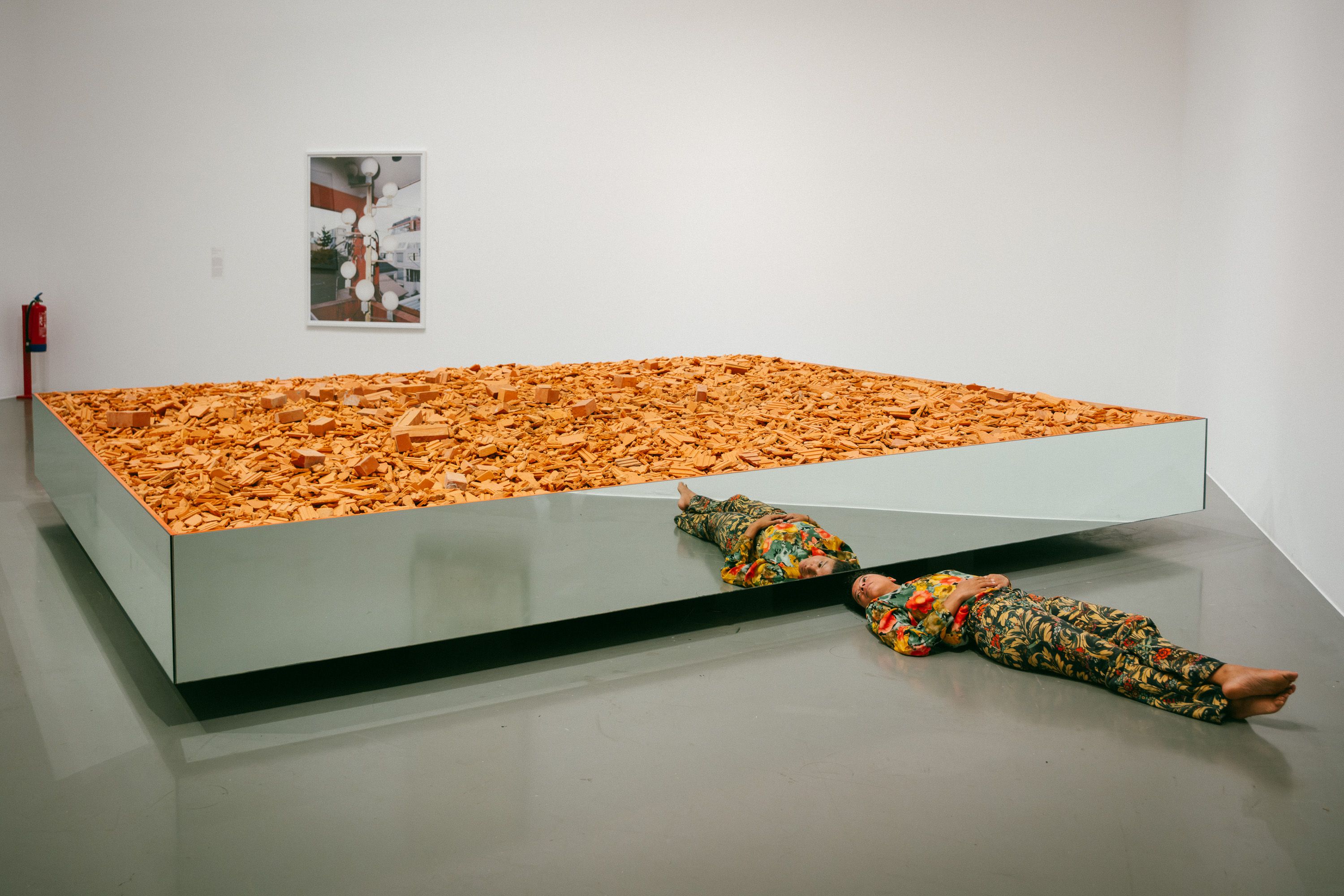 A person wearing a colorful floral suit lies on their back on the floor of a museum, parallel to a large, rectangular mirrored platform filled with terracotta bricks and rubble. Their reflection is visible in the mirrored base of the structure. In the background, a framed artwork hangs on a white wall. - Diana Mendes