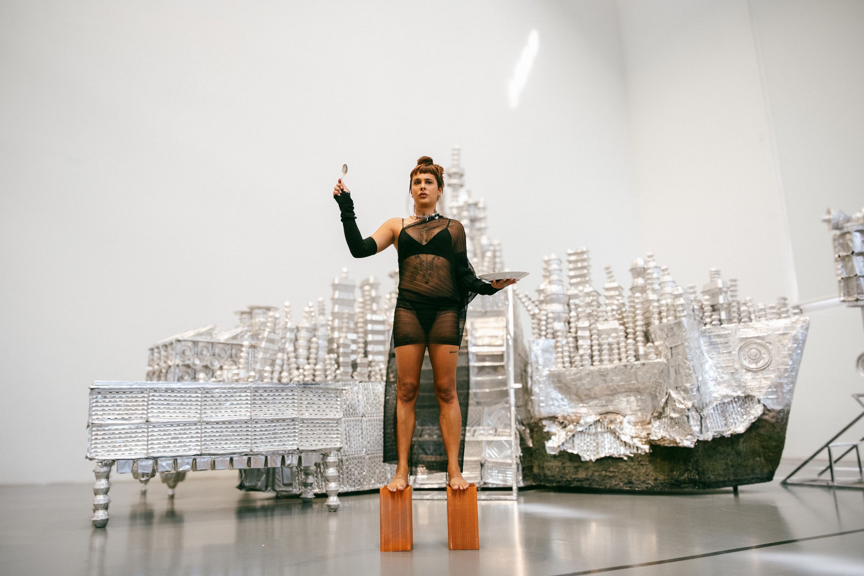 Wide shot of an artist balancing on two wooden blocks inside a spacious art gallery. She wears a sheer black outfit, holding a raised spoon and a plate. Behind her is a large silver art installation made of aluminum containers resembling buildings and a boat. - Diana Mendes