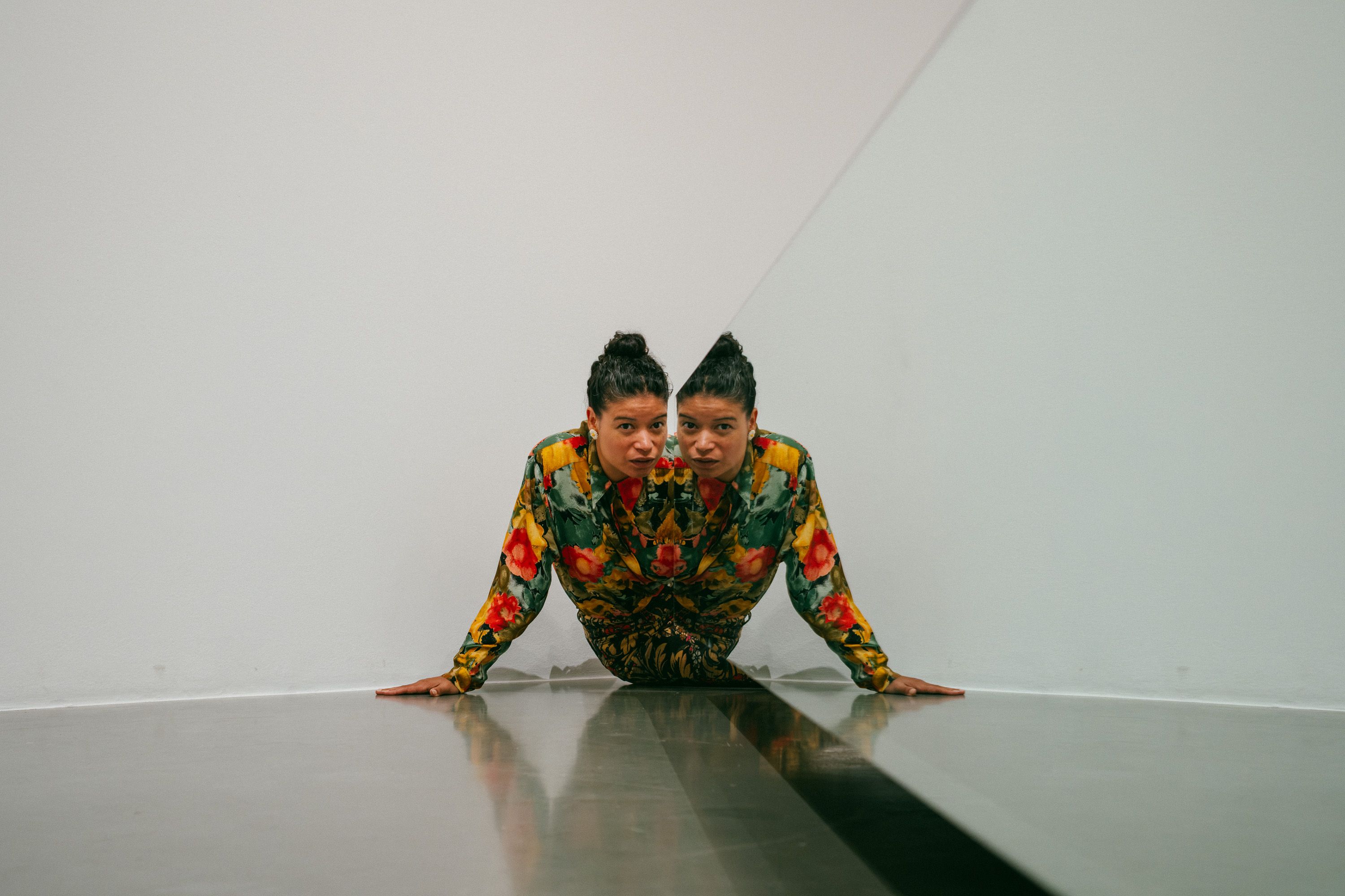 A person in a floral outfit with her hair in a bun leans against the corner of a mirrored wall, creating a symmetrical optical illusion of mirrored bodies. She rests her hands on the gray floor, looking directly at the camera. - Diana Mendes