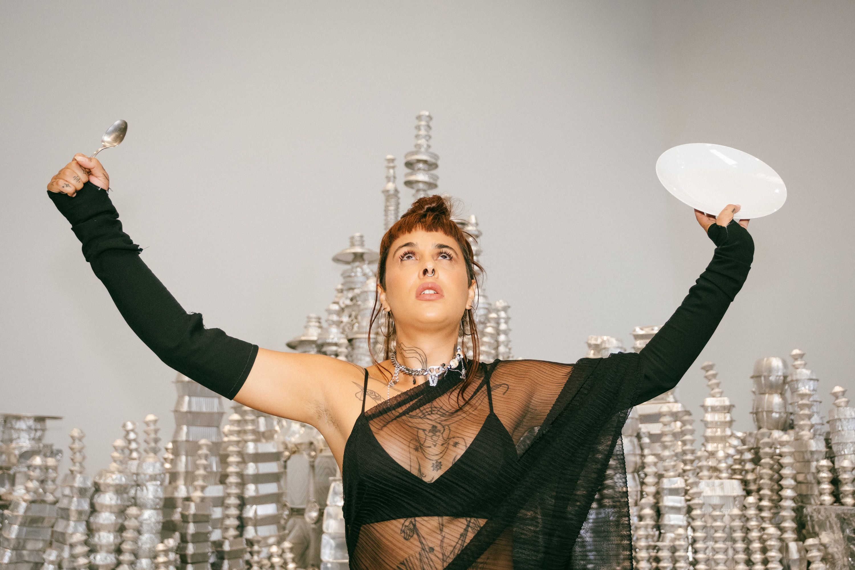 Close-up of a performer with short brown hair and bangs, wearing a sheer black dress and long arm sleeves. She looks upward with raised arms in a "V" shape, holding a metal spoon in her right hand and a white plate in her left hand. Stacked, shimmering silver structures are visible in the background. - Diana Mendes