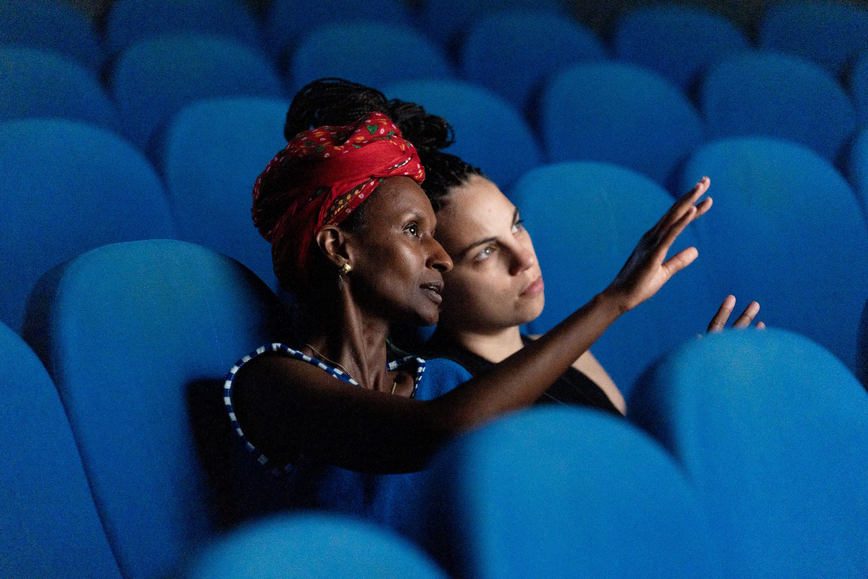 © Pietro Bertora Medium shot of two people seated in blue cinema chairs. The woman in the foreground wears a patterned red headscarf and a blue top, pointing with an raised hand as she speaks to the person next to her, who listens intently. - Pietro Bertora