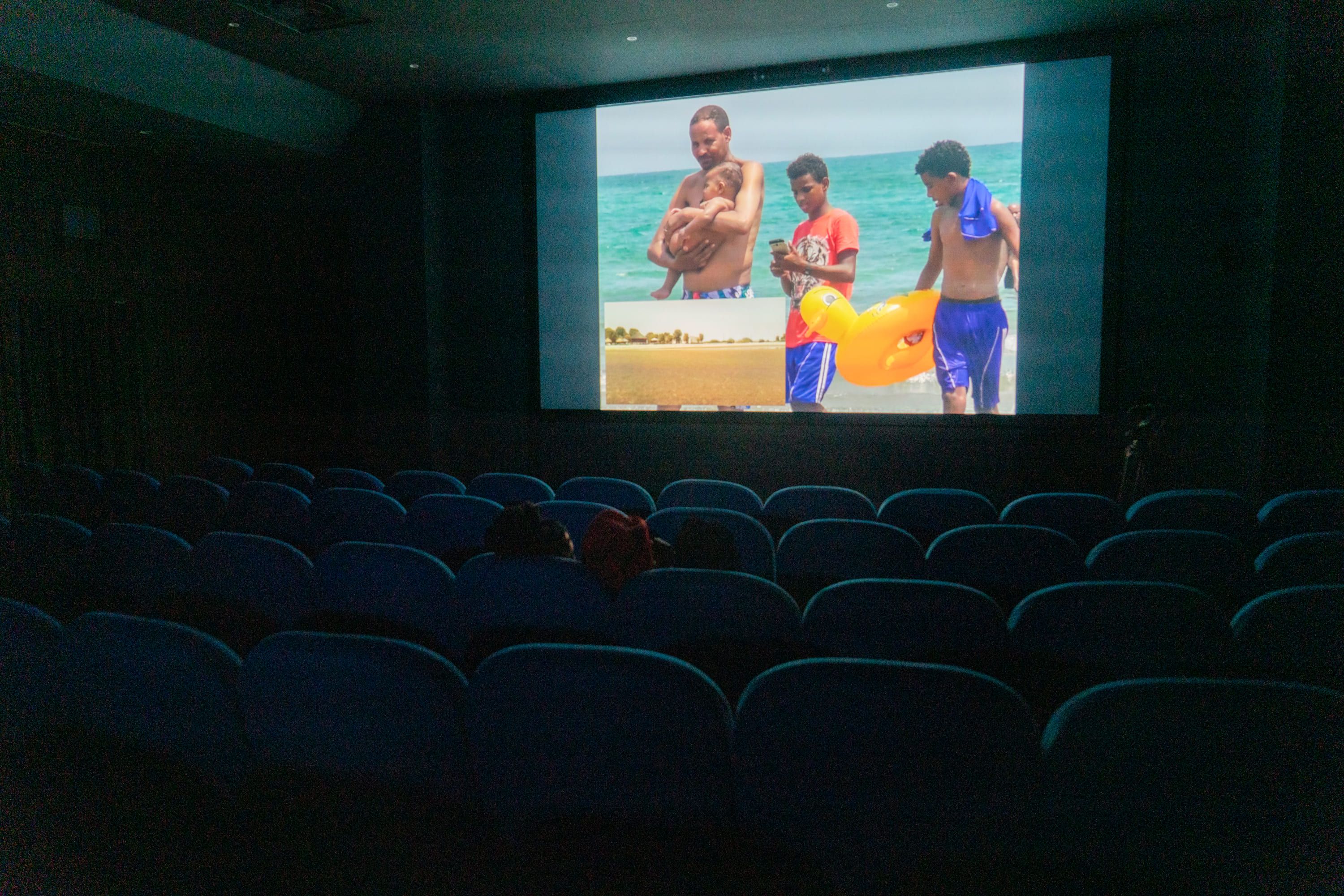 © Pietro Bertora Wide shot of a dark cinema auditorium with rows of mostly empty blue seats. A colour photograph of several people on a beach by the sea is projected onto the screen at the front. - Pietro Bertora