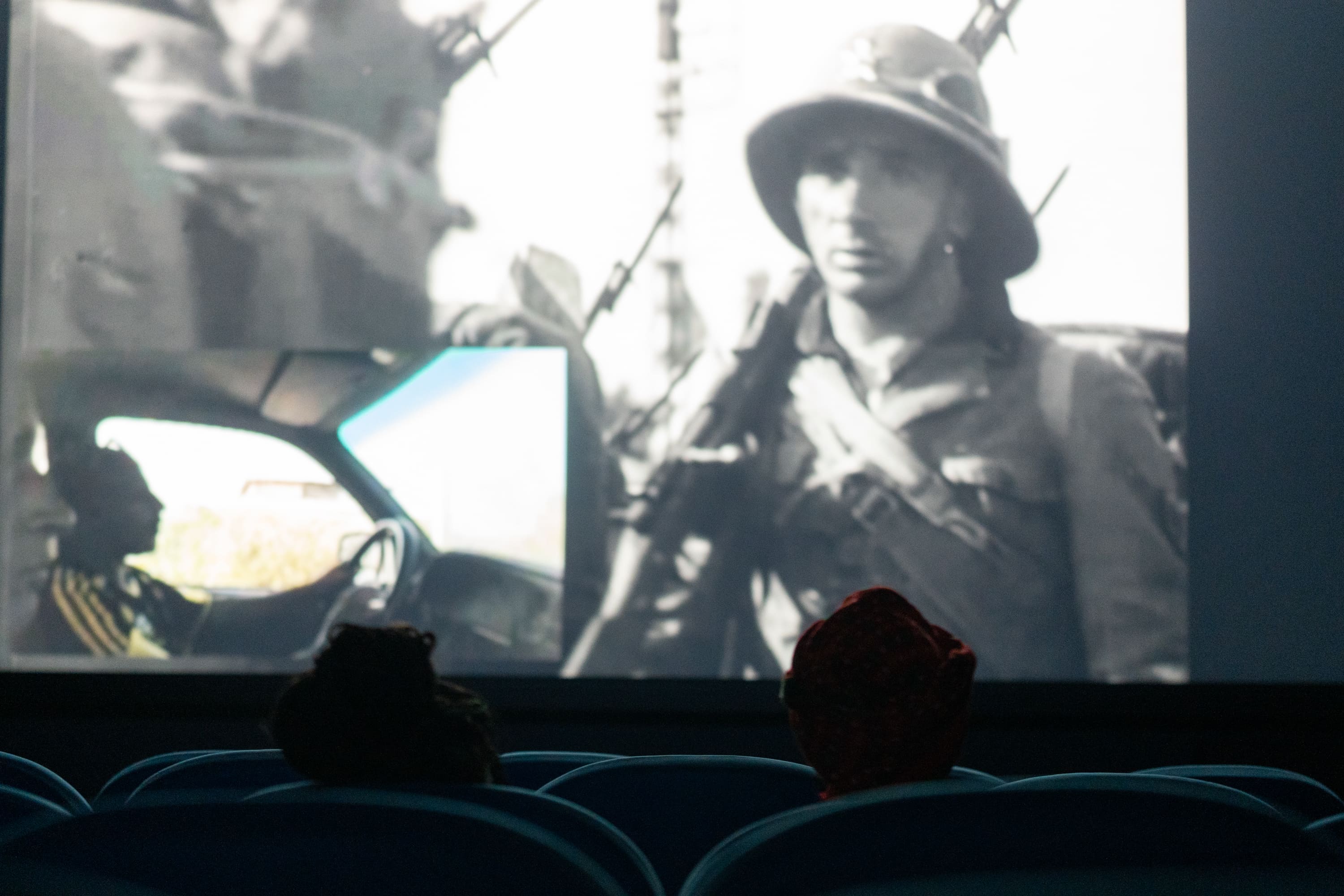 © Pietro Bertora View from the back of a dark auditorium showing the silhouettes of two audience members in cinema seats, watching a film projection. The screen displays a historical black-and-white image of a helmeted soldier with a smaller picture-in-picture insert of someone driving. - Pietro Bertora