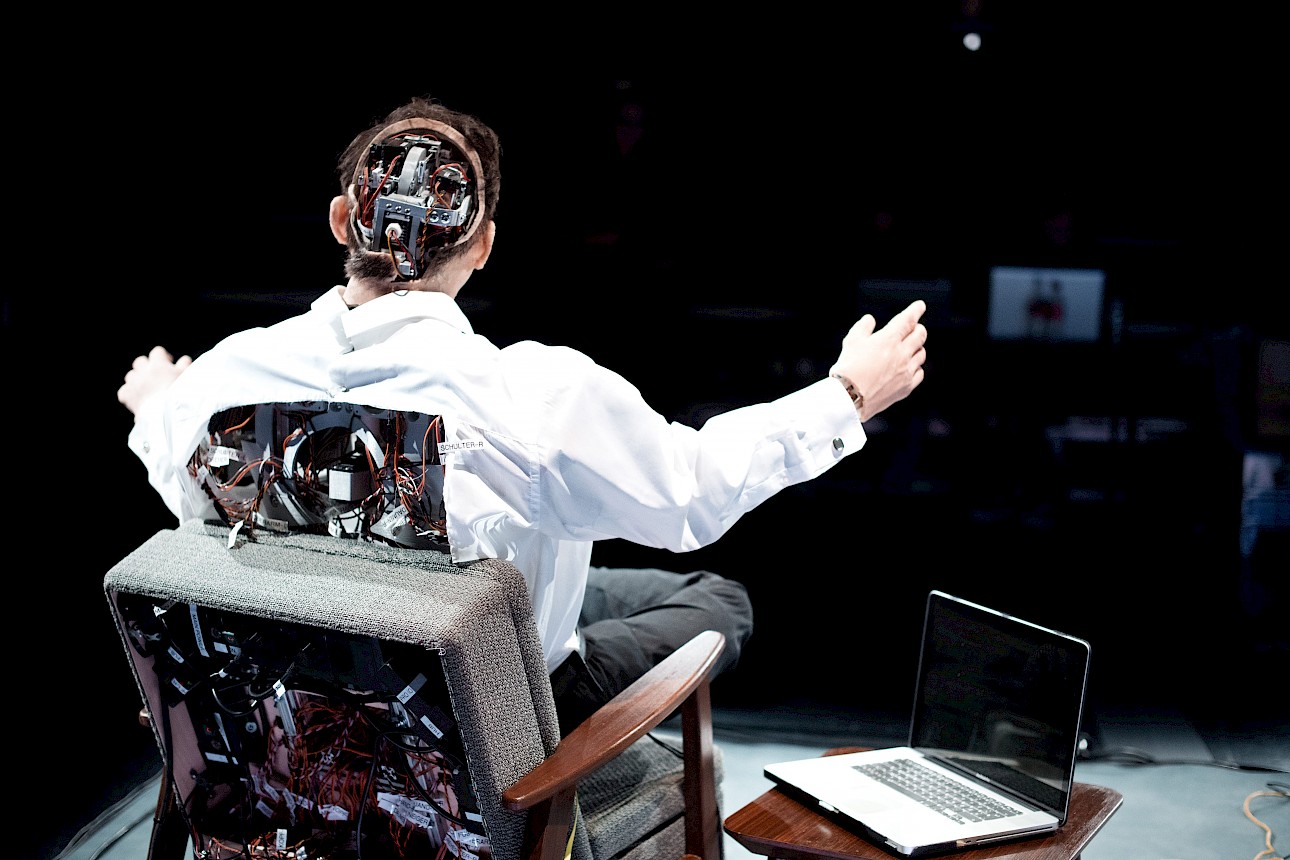 ALKANTARA - Rear view of a figure in a white shirt seated in an armchair with open arms on a dark stage. The back of its head and torso are open, exposing wires, circuits, and mechanical parts. An open laptop rests on a side table nearby. - ©Gabriela Neeb
