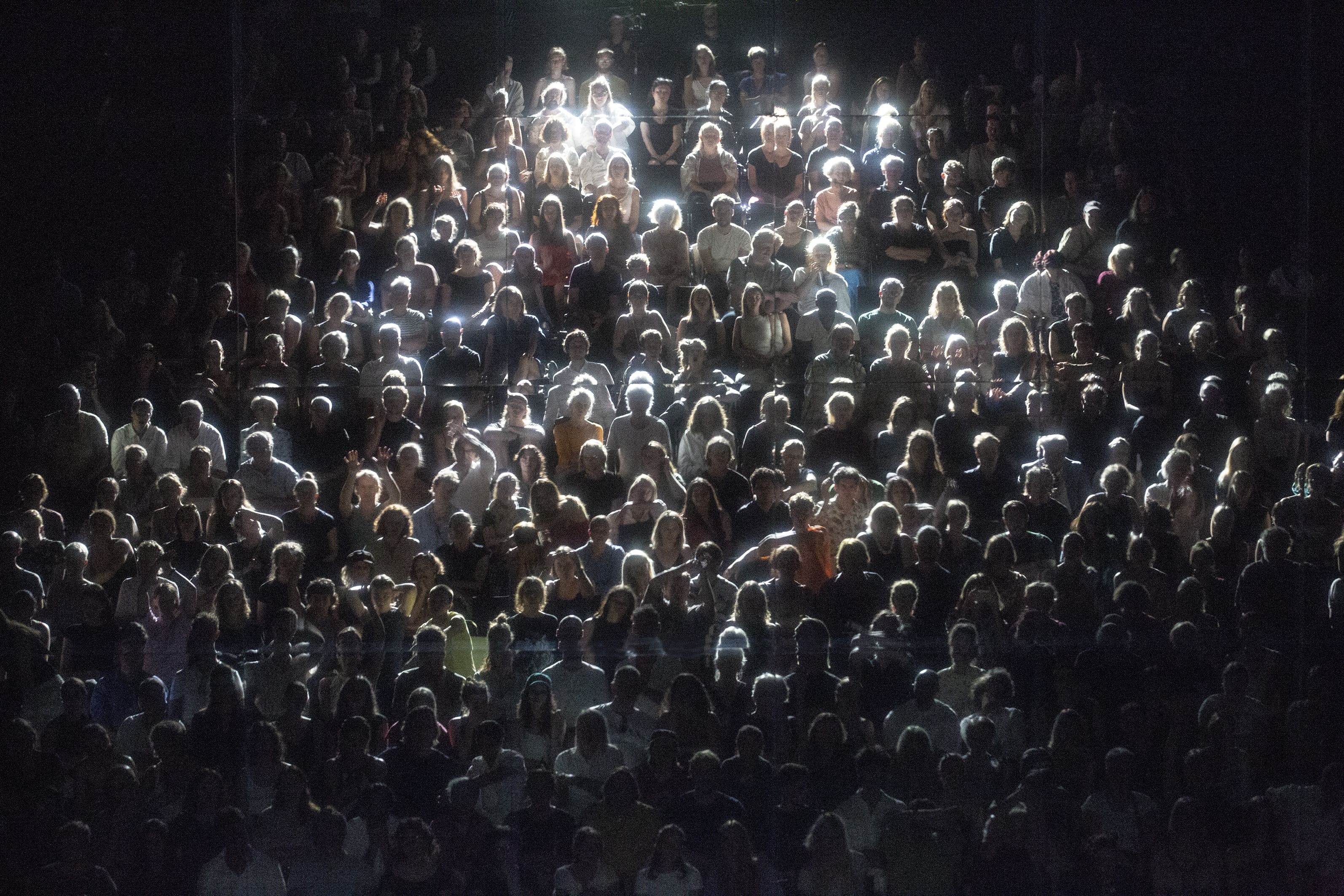 © Bernd Uhlig A large, dense audience seated in a dark auditorium. Bright overhead lighting catches the tops of people's heads and shoulders, creating glowing, high-contrast outlines against their faces and bodies in the darkness. - Bernd Uhlig