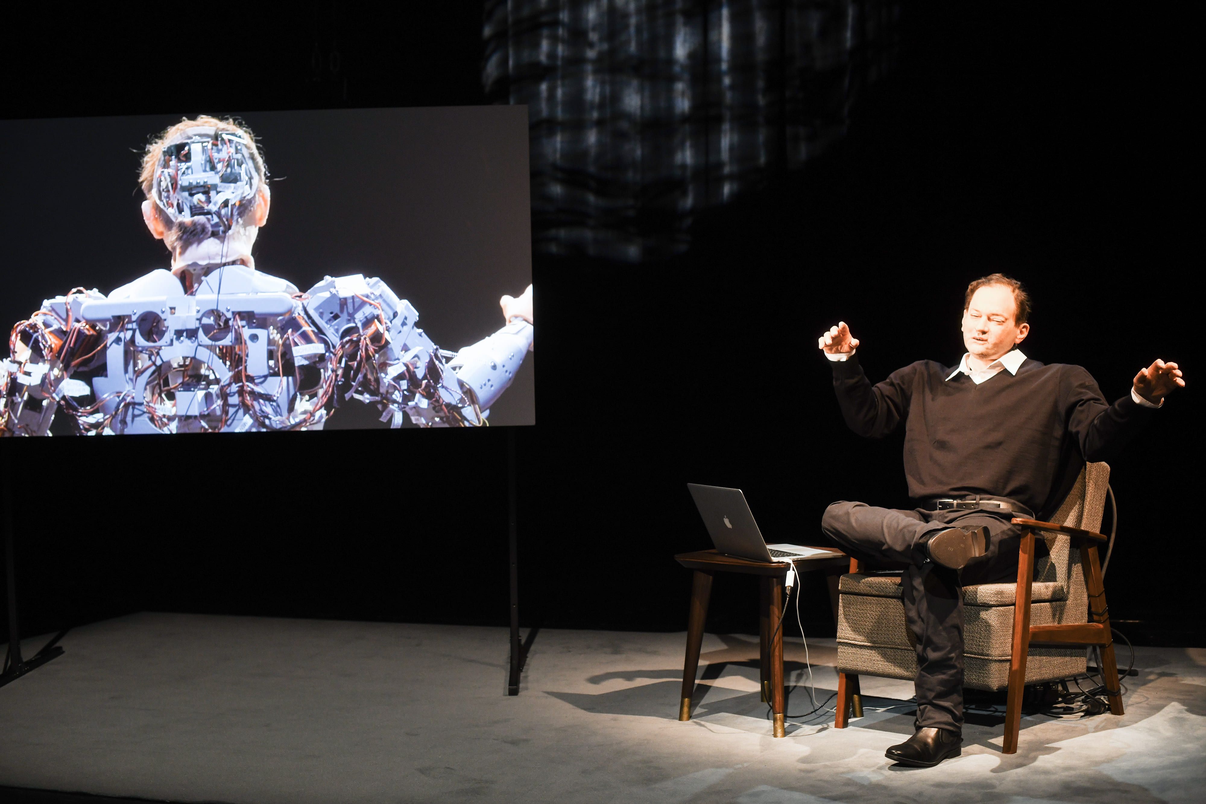 © Gabriela Neeb On a dark stage, a man in a black jumper sits cross-legged in an armchair with his arms raised, next to a laptop on a small table. On the left, a screen displays a close-up image of the back and mechanical head of a humanoid robot. - Gabriela Neeb