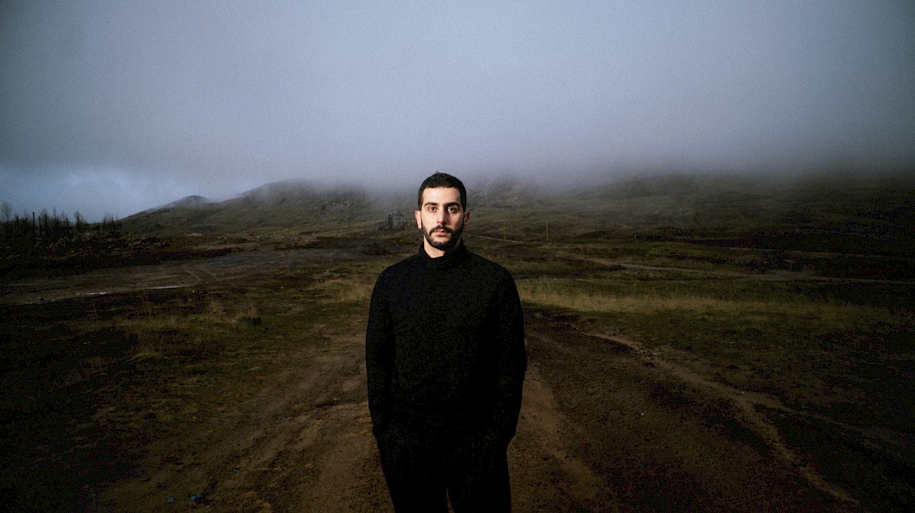 ALKANTARA - Ali Chahrour portrait. Medium portrait of a man with a short beard and dark hair, wearing a black roll-neck jumper and black trousers, standing on a dirt path facing forward. In the background, a hilly rural landscape unfolds under a cloudy, misty sky. - ©Lea Skayem