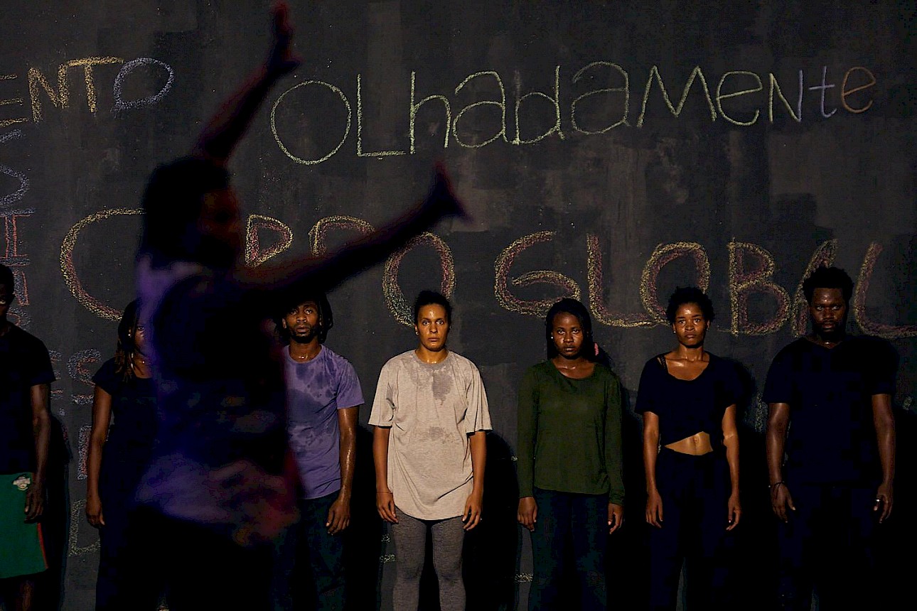 ALKANTARA - In the blurred foreground, a figure gestures with raised arms towards a line of six performers in a rehearsal space. The performers stand side by side with focused expressions, wearing casual sportswear, in front of a dark wall covered in handwritten chalk text. - ©Mariano Silva