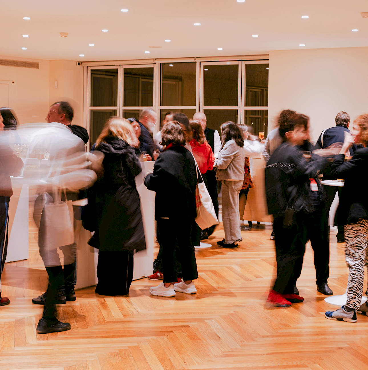 ALKANTARA - Guests mingle and converse at a social gathering inside a brightly lit room with herringbone wooden flooring and recessed ceiling lights. Some figures appear slightly blurred due to motion blur. Large glass windows are visible in the background. - ©Beatriz Pequeno