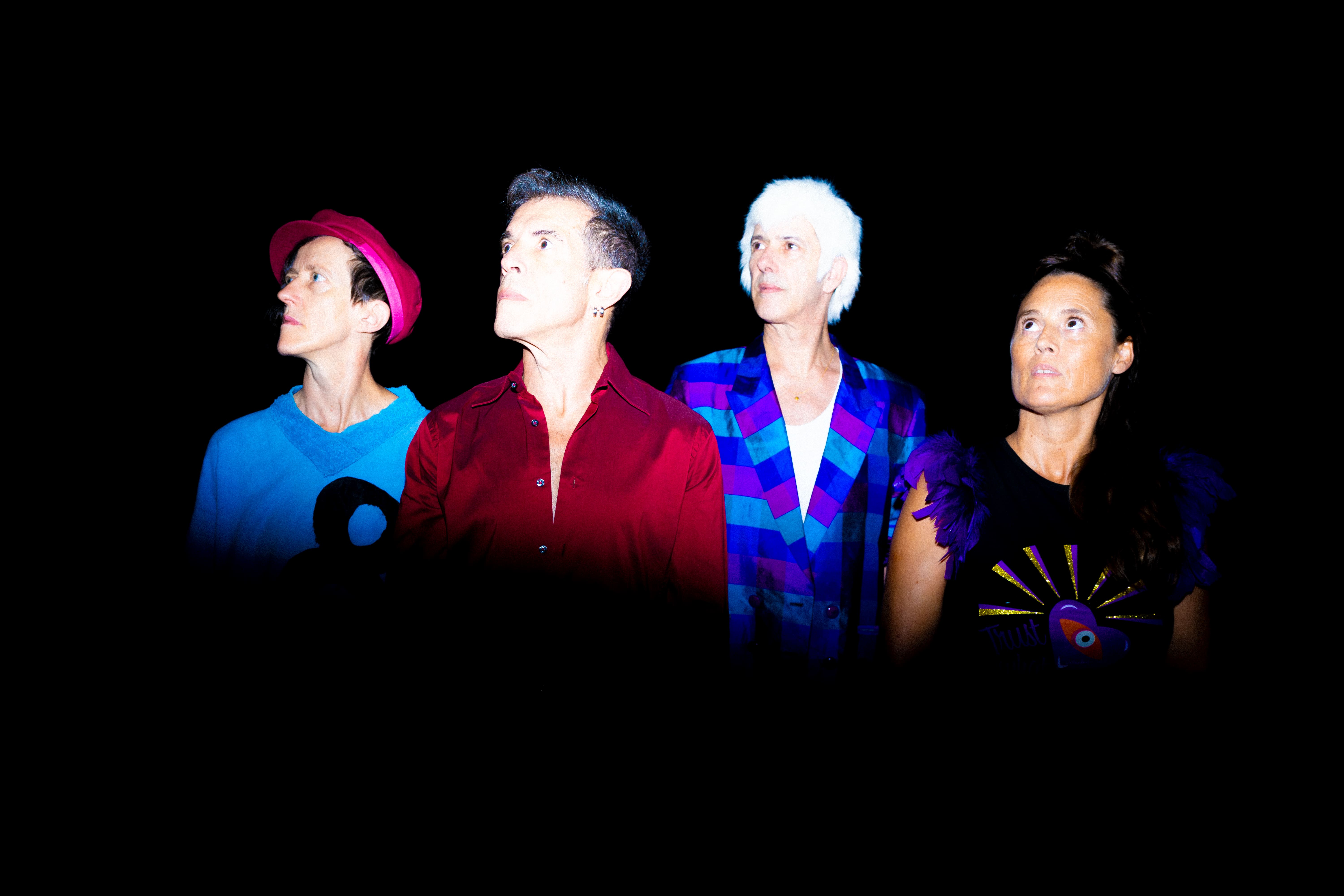 © Alípio Padilha Four people pose lined up against a solid black background under flash lighting, all looking upwards to the left with a contemplative expression. The person on the left wears a bright pink hat and a light blue t-shirt; the second wears a burgundy shirt; the third wears a short white wig and a blue and purple geometric blazer; the person on the far right wears a black top with illustrated details. - Alípio Padilha