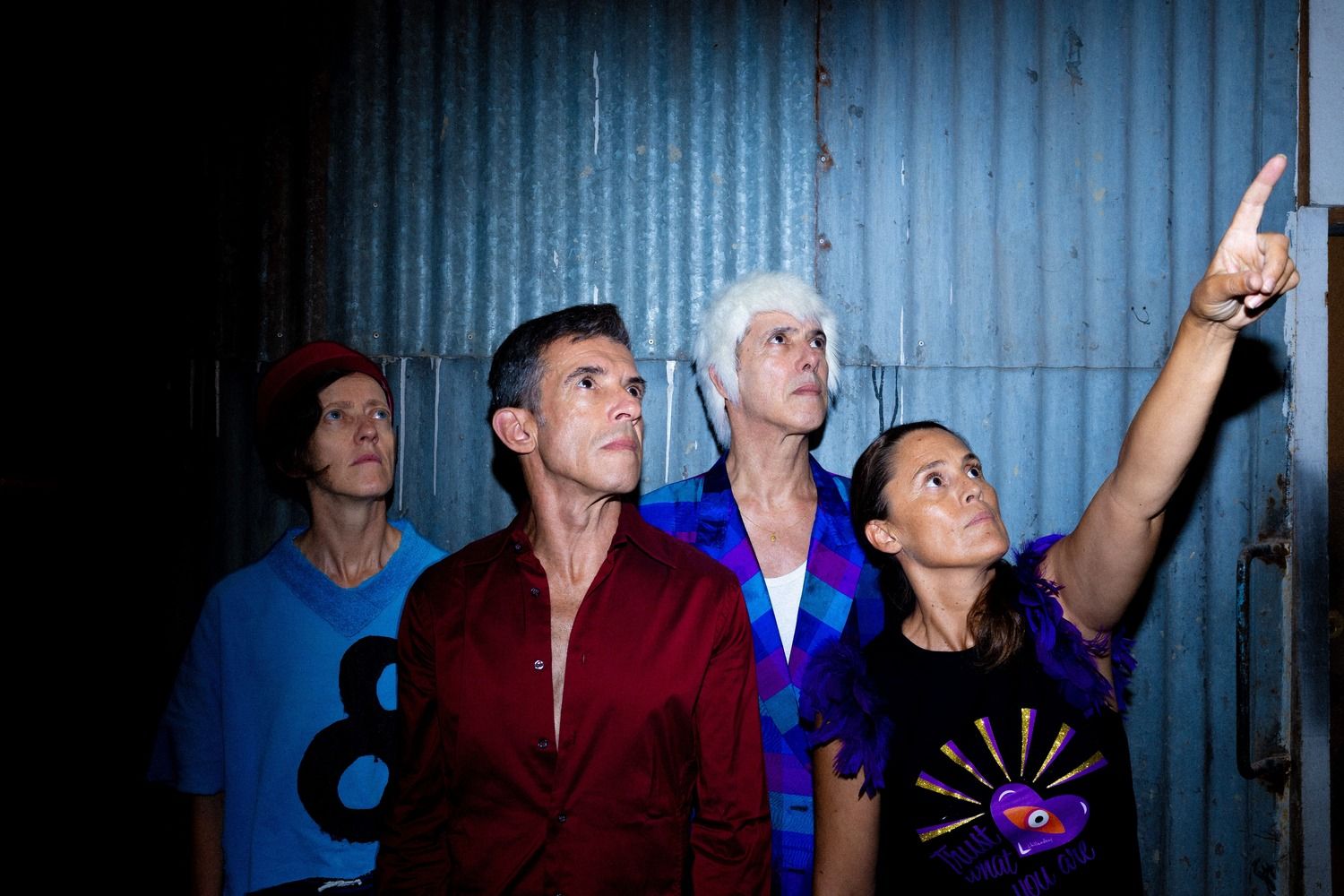 © Alípio Padilha Four people pose in front of a corrugated metal wall under flash lighting. The person on the right wears a black top with purple feathers on the shoulders and points upwards with her index finger towards the top right, while the other three people look intently in that direction. - Alípio Padilha