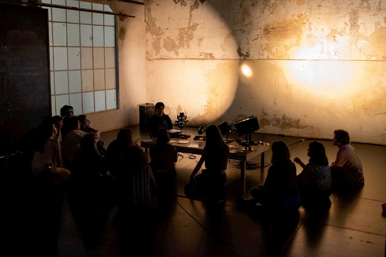 ALKANTARA - A group of people sits in a semicircle on the dark floor of an industrial-style room with textured, distressed plaster walls and a tall grid window in the background. In the centre, a long table holds various spotlights and lighting equipment, projecting two circles of warm, yellowish light onto the back wall. - ©Juca Schindler