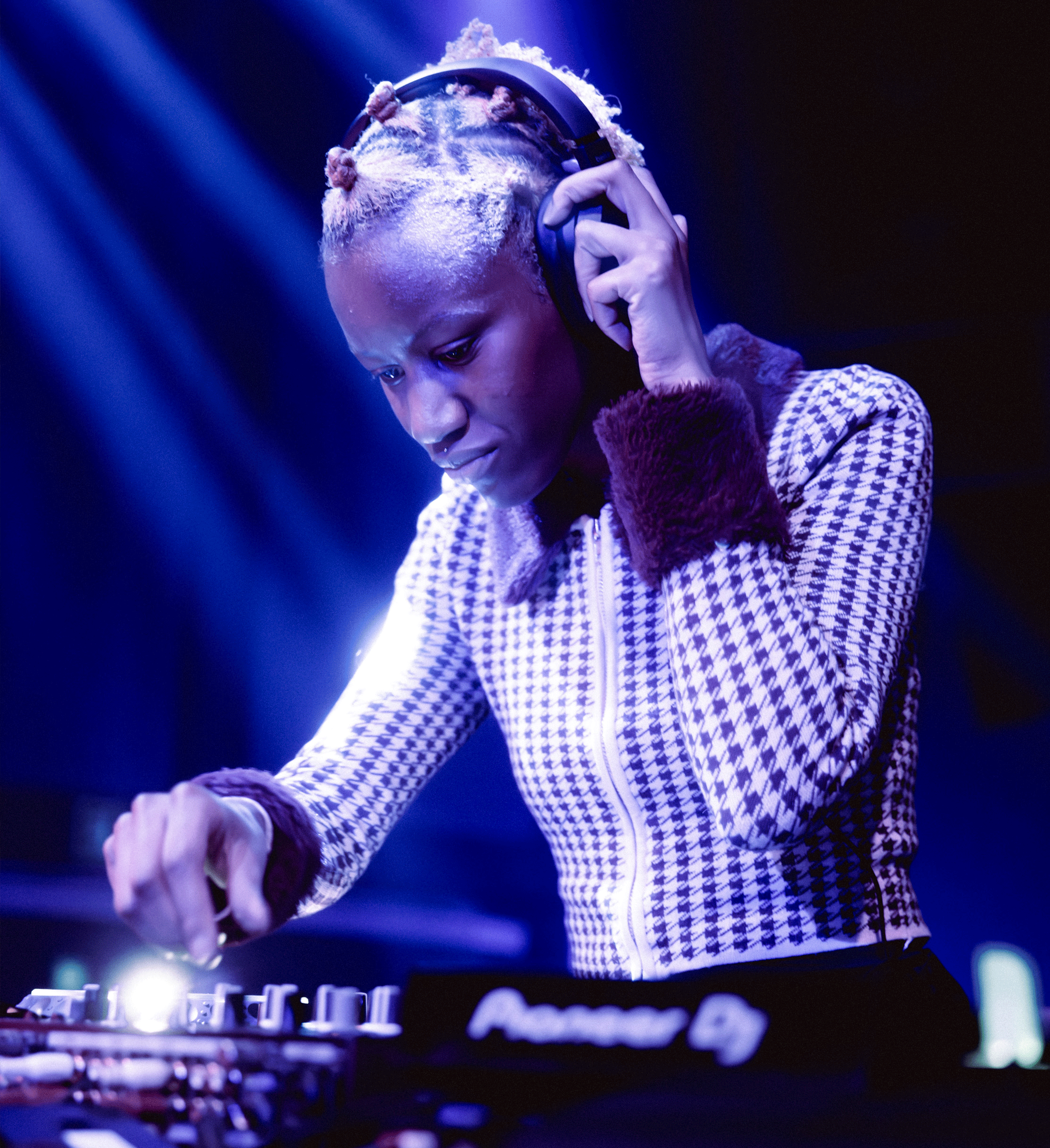 © tomicornio Plano de uma pessoa DJ negra de cabelo descolorido apanhado em pequenos coques, concentrada a misturar música numa mesa de som sob iluminação azul. Segura auscultadores numa das orelhas e veste um casaco com padrão houndstooth em tons escuros e claros com pormenores em veludo nas mangas e gola. - tomicornio