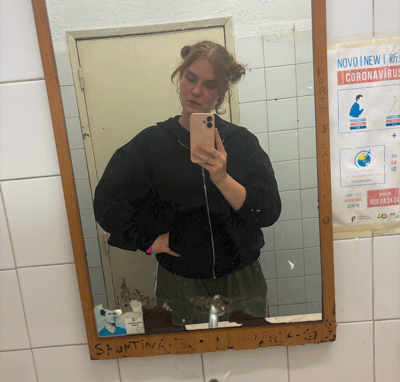 ALKANTARA - Selfie de Inês Coutinho tirada em frente ao espelho de uma casa de banho com azulejos brancos. Tem o cabelo apanhado em dois ganchos/coques laterais, veste um casaco com capucho preto e segura um telemóvel rosa claro, posando com uma mão na cintura e os olhos semicerrados. Na moldura do espelho e na parede ao lado, veem-se autocolantes, inscrições e um cartaz informativo. - ©Inês Coutinho