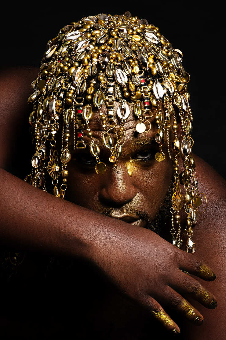 Retrato de DIDI. Plano aproximado de uma pessoa negra com o rosto decorado com maquilhagem dourada e um toucado elaborado de búzios e pendentes dourados. - Feeh Garcia
