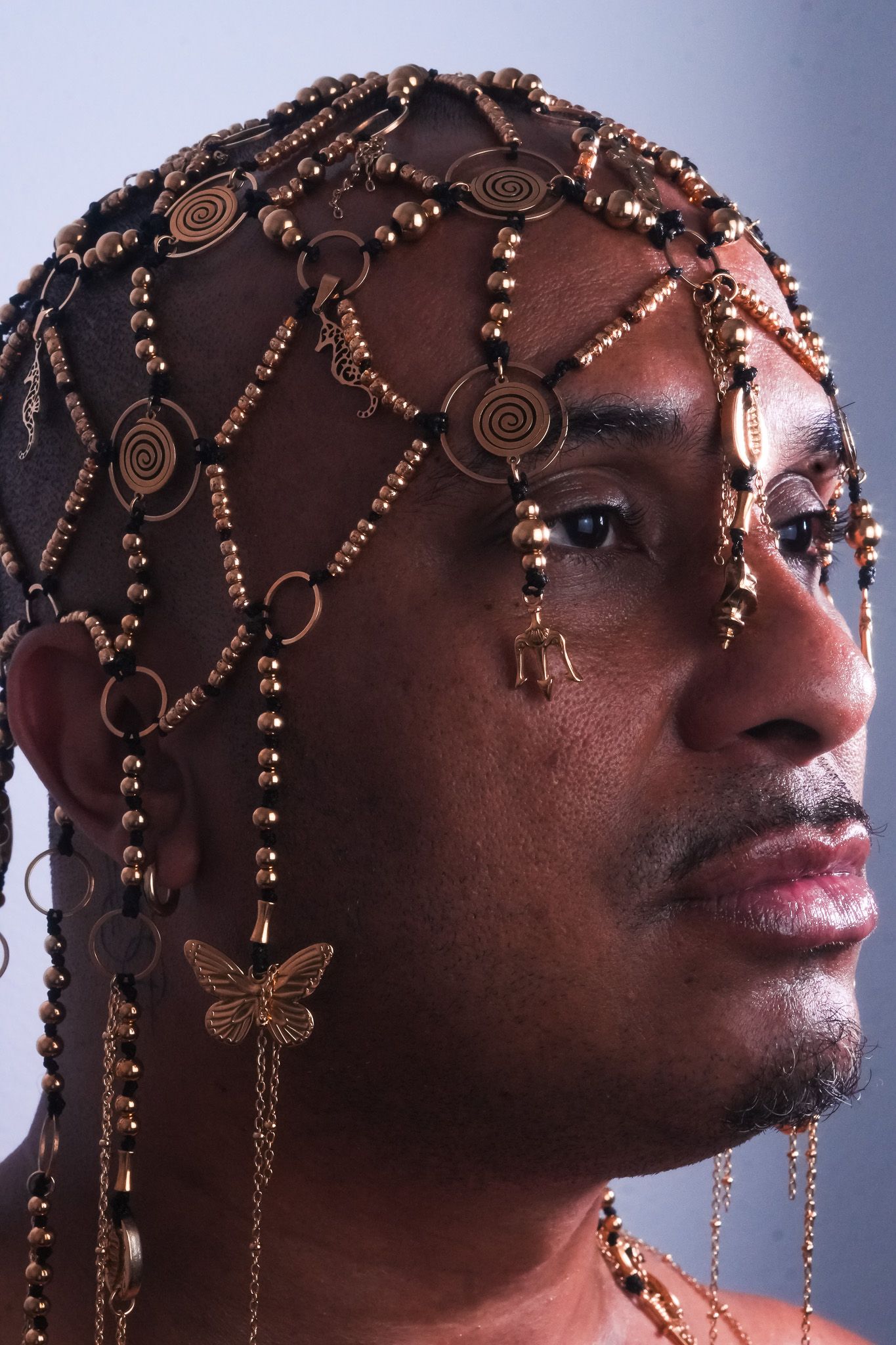 Retrato em perfil de Pedro Barbosa. Uma pessoa negra a usar uma peça de cabeça dourada e intrincada com contas, correntes e pendentes, incluindo uma borboleta. - Joni Ricos