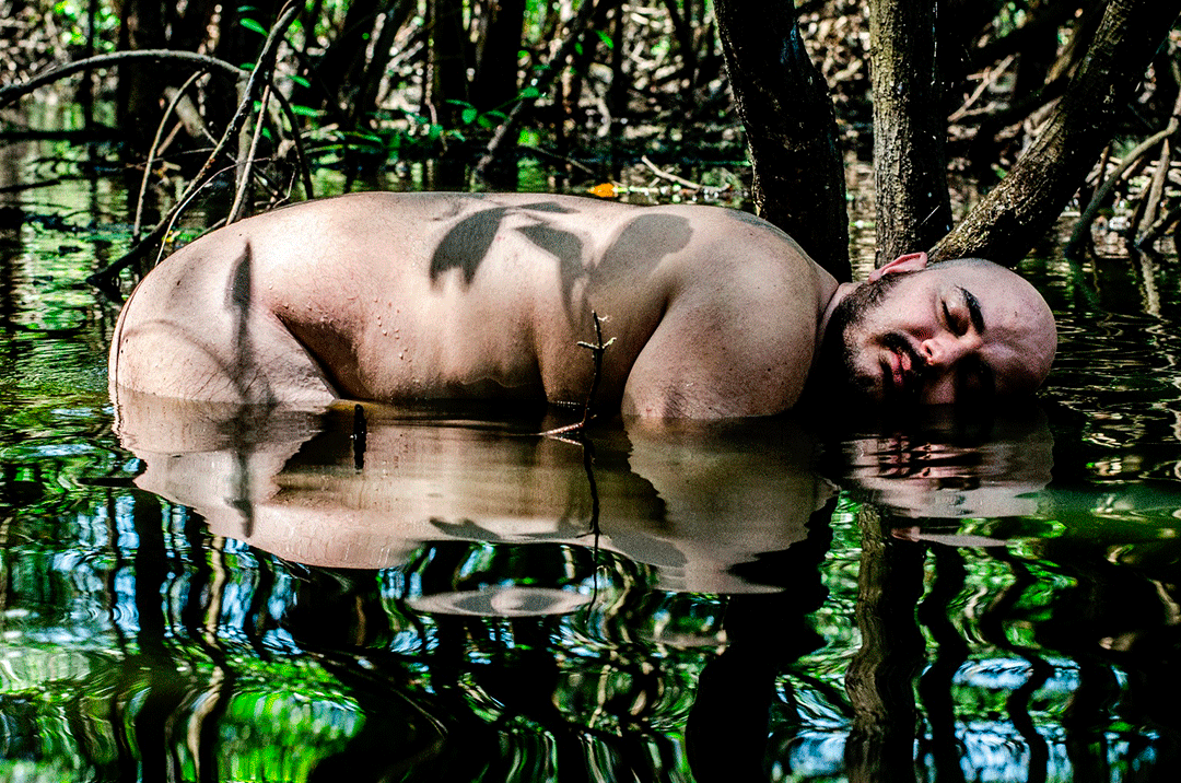Uma pessoa de tronco nu e cabeça rapada a flutuar na água de um mangueiral ou pântano denso, rodeada por árvores e vegetação refletidas na superfície da água. - Luiz Alves