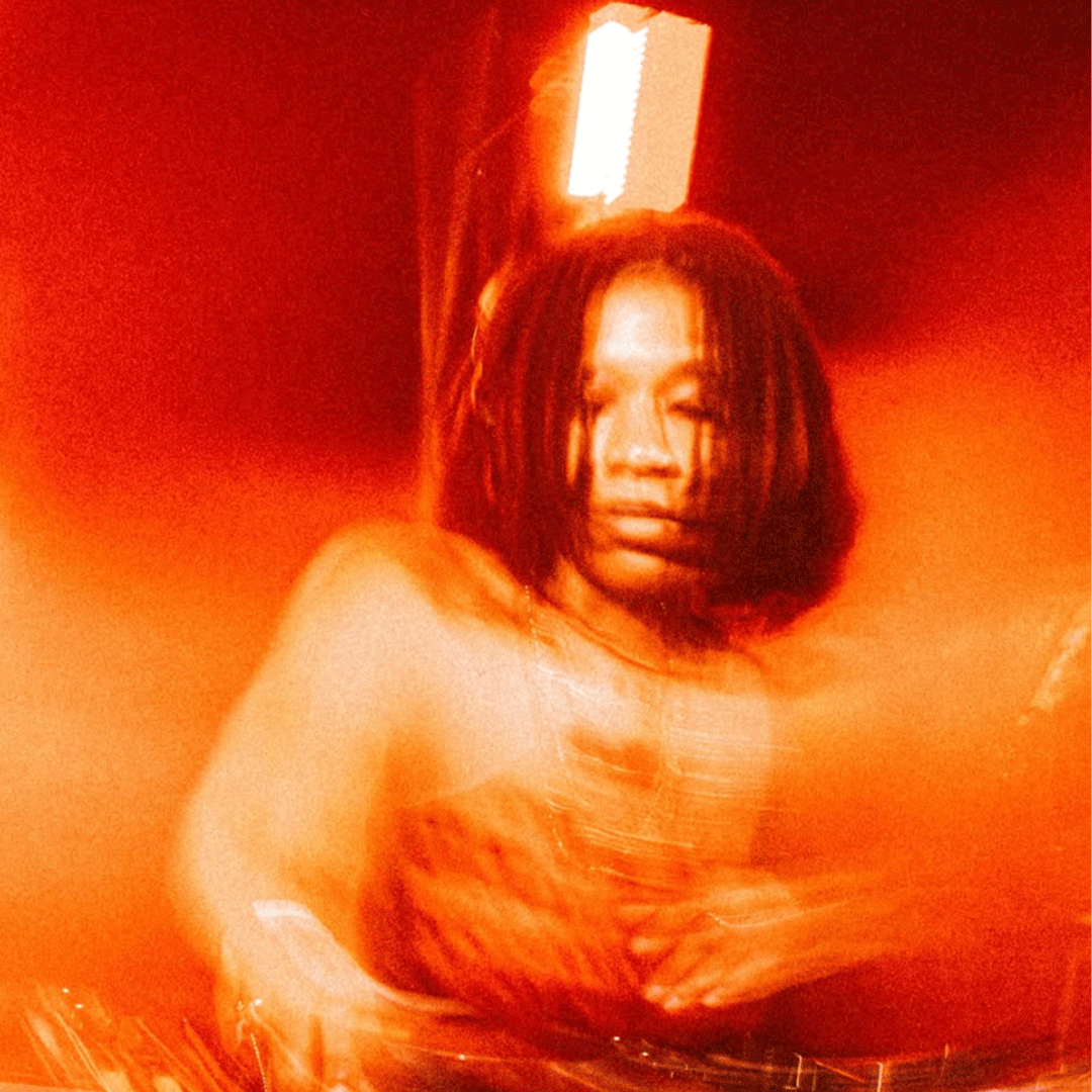 Retrato com efeito de movimento e tonalidade vermelha e laranja vibrante, mostrando uma pessoa de dreadlocks curtos a atuar como DJ atrás de uma mesa de som com iluminação quente ao fundo.