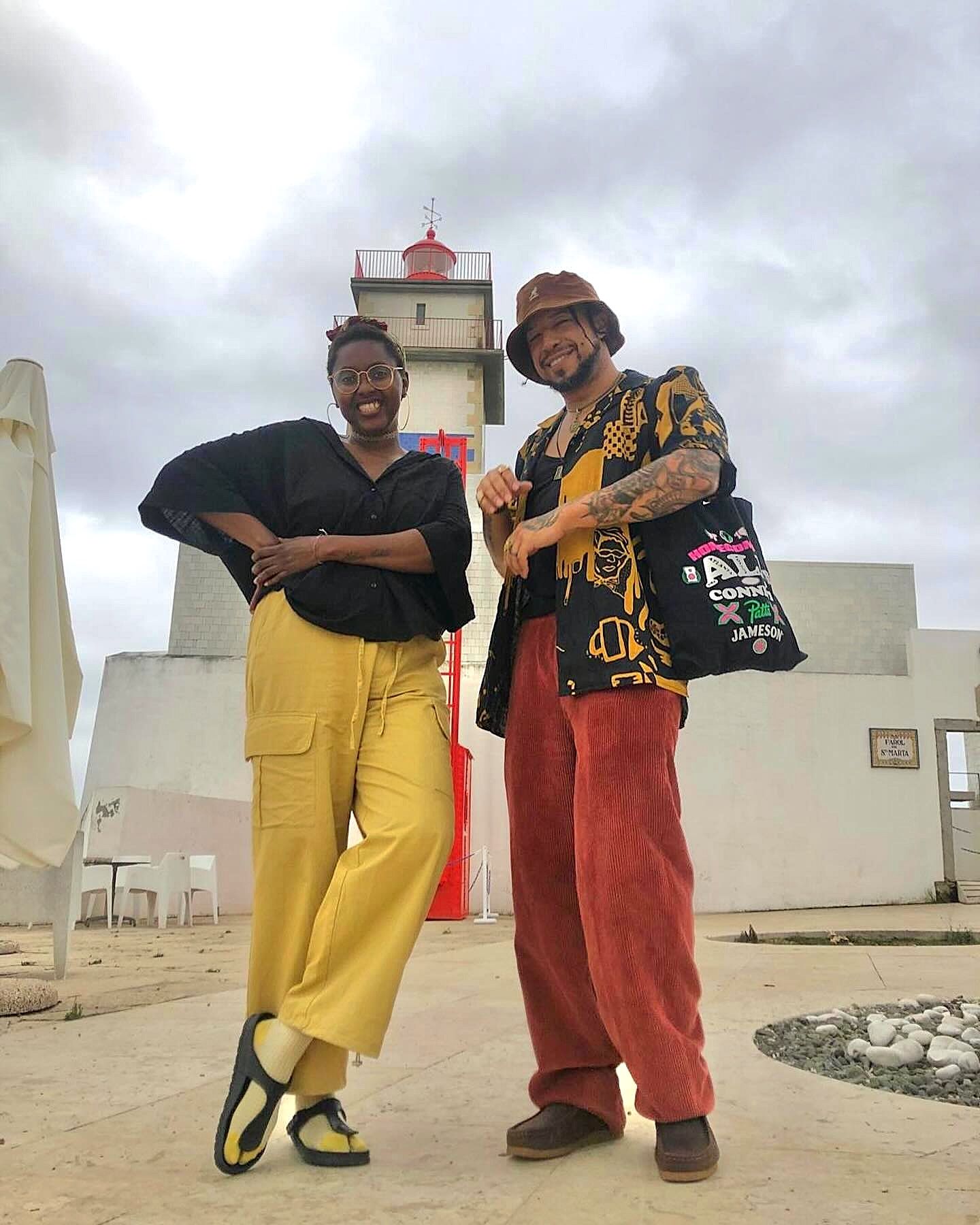 Duas pessoas posam sorridentes ao ar livre em frente a um farol sob um céu nublado. A pessoa da esquerda veste uma blusa preta, calças amarelas cargo e óculos redondos, posando com a mão na cintura. A pessoa da direita usa um chapéu bucket castanho, camisa estampada em preto e amarelo sobreposta a uma t-shirt preta, calças largas de veludo cinto-de-vassoura vermelho/tijolo e transporta um saco de pano ao ombro.