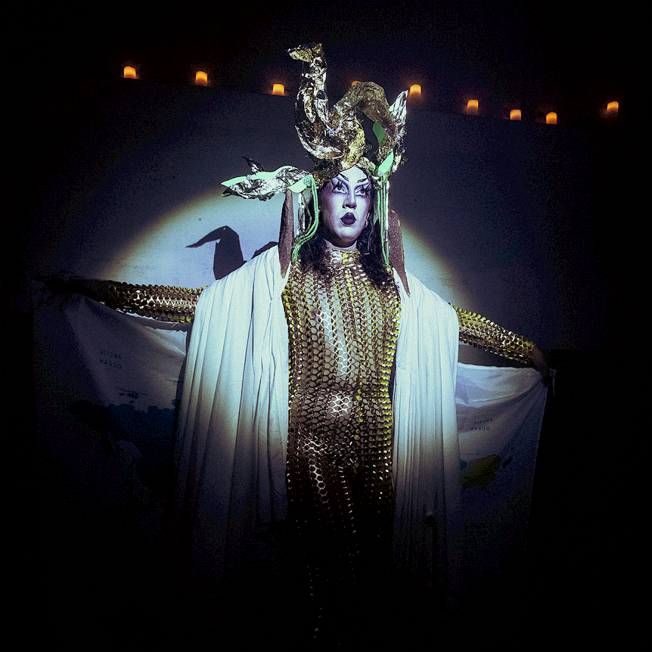 ALKANTARA - Pessoa em performance vestida com um fato justo em malha dourada de lantejoulas e uma capa branca drapeada, posando de braços abertos num palco escuro sob um foco de luz circular. Destaca-se um adereço dourado elaborado na cabeça e uma maquilhagem dramática, segurando nas mãos o que parece ser um mapa-múndi impresso em tecido. - ©tomi cornio