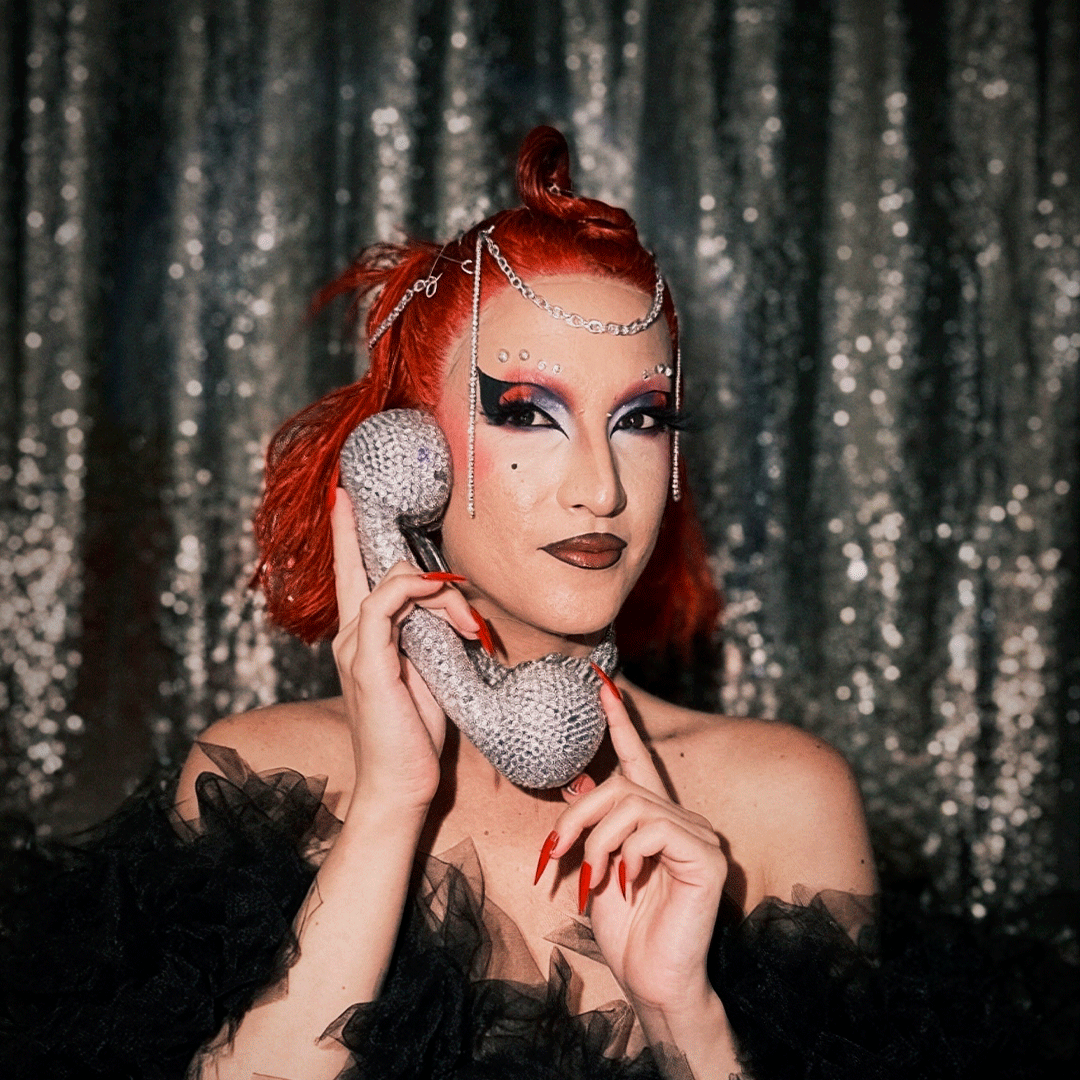 Retrato em plano médio de uma pessoa em performance de cabelo ruivo vivo apanhado no topo e maquilhagem drag estilizada, a segurar ao ouvido um auscultador de telefone vintage prateado coberto de brilhantes. Veste um fato/vestido preto com folhos de tule, com unhas compridas vermelhas e correntes prateadas a embelezar o penteado, em frente a um fundo reluzente de lantejoulas prateadas.