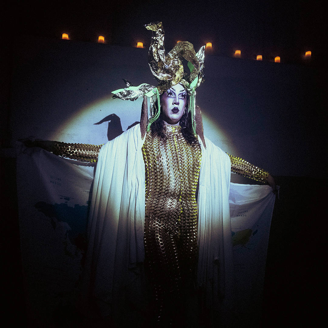 © tomi cornio Pessoa em performance vestida com um fato justo em malha dourada de lantejoulas e uma capa branca drapeada, posando de braços abertos num palco escuro sob um foco de luz circular. Destaca-se um adereço dourado elaborado na cabeça e uma maquilhagem dramática, segurando nas mãos o que parece ser um mapa-múndi impresso em tecido. - tomi cornio