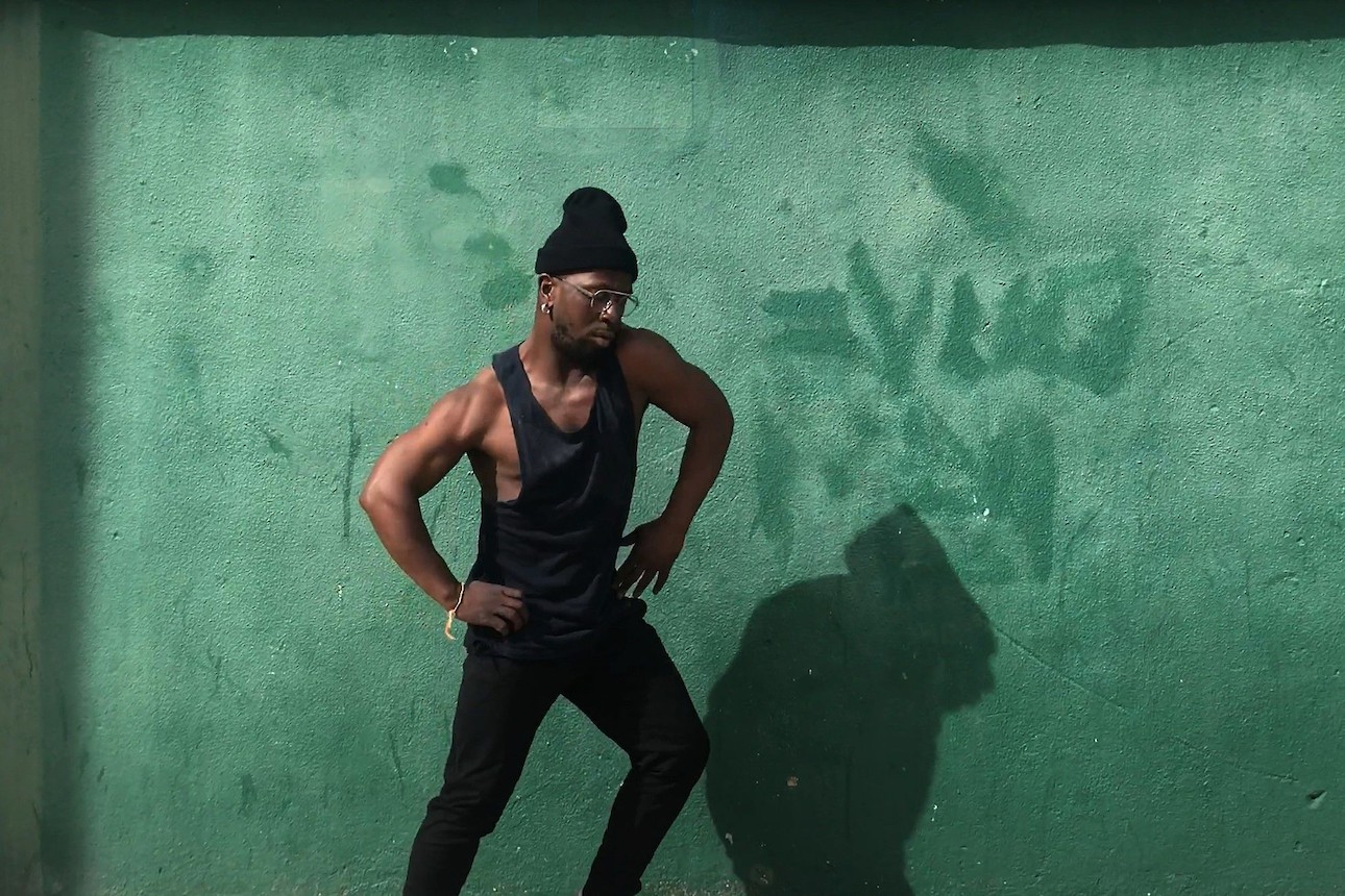 ALKANARA - Imagem de fundo é uma parede verde, mostra um homem adulto no centro com as mãos na cintura e pernas arqueadas em movimento. Homem veste calças prestas, blusa de alças preta, gorro preto e óculos escuros. - ©Sofia Berberan